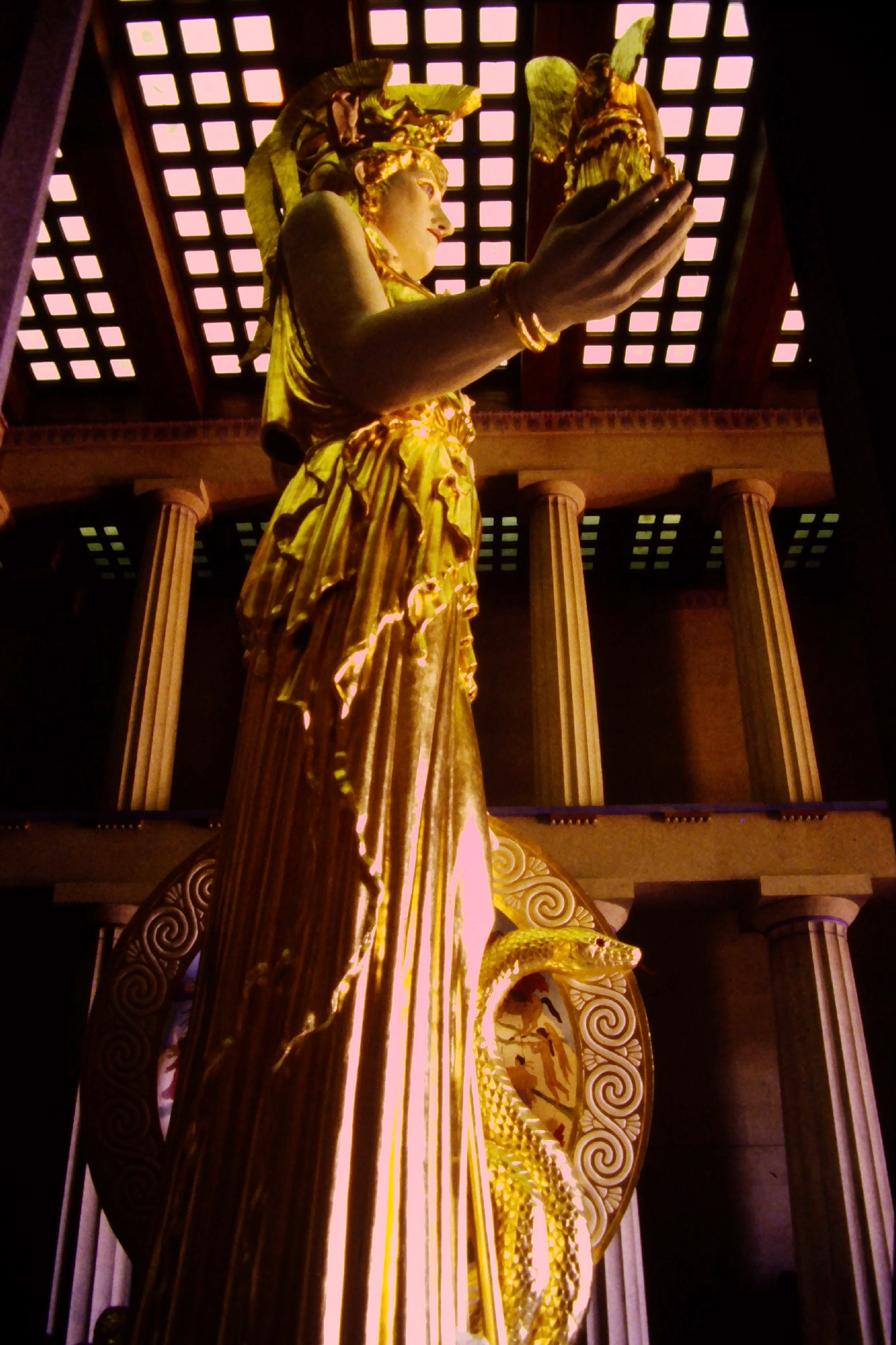 Close-up of a golden statue of a historical figure, possibly a Greek or Roman deity or hero, with ornate drapery and detailed craftsmanship, set against a background of wooden columns and a patterned ceiling with interior lighting. Athena Parthenos