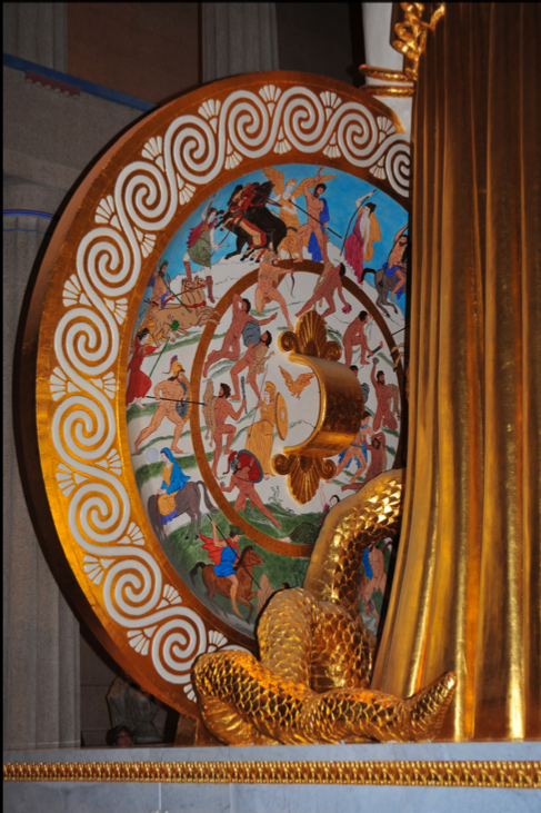 Decorative circular artwork with intricate patterns, depicting mythological or historical scenes with figures, horses, and a golden serpent, partly covered by a golden curtain on the right side. Athena Parthenos Shield. 