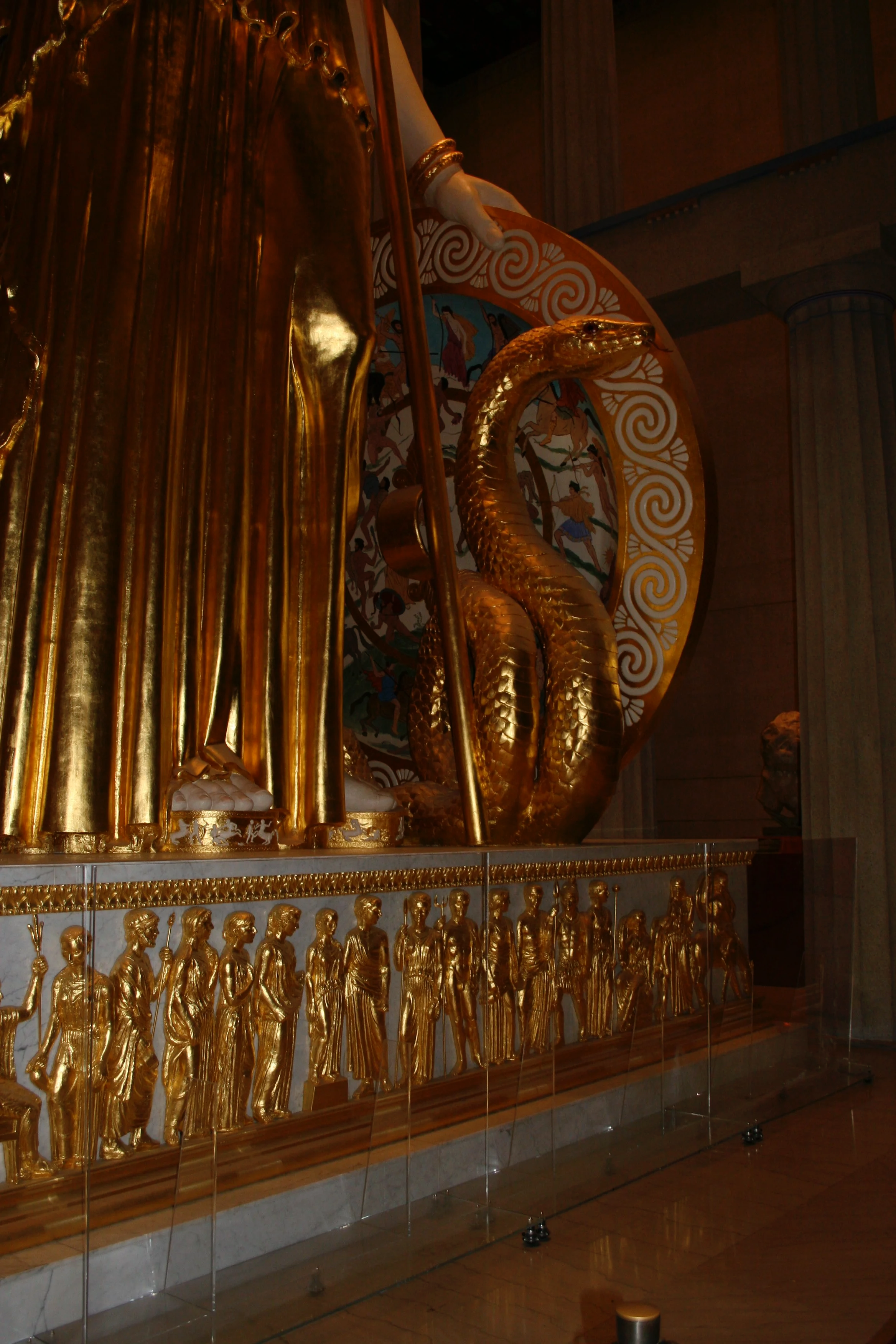 A golden sculpture of a snake with a detailed head, part of a larger ornate display that includes gold figures of people and decorative circular patterns in the background. Athena Parthenos in Parthenon Museum Nashville. 
