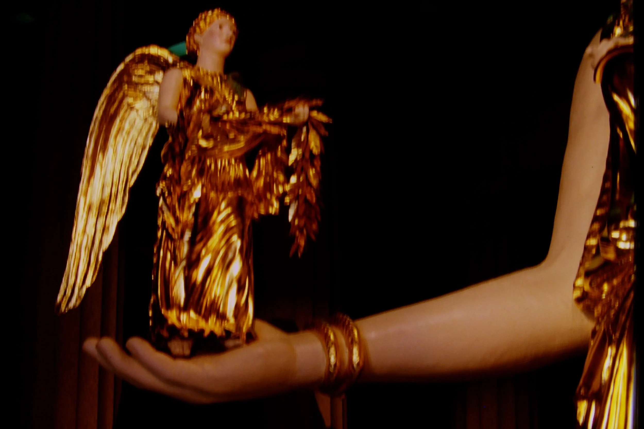 A person holding Nike in her hand with gold and orange patterned clothing and jewelry, standing against a dark background. Athena Parthenos in Parthenon Museum Nashville. 