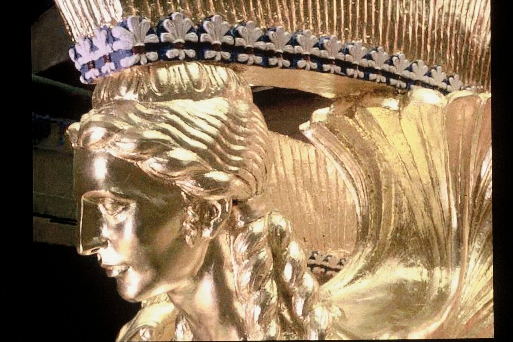 A close-up of a metallic or gold-colored ornate helmet, featuring a figure with a detailed face, flowing hair, and a solemn expression, with decorative elements and a crown or headdress above. Athena Parthenos in Parthenon Museum Nashville.