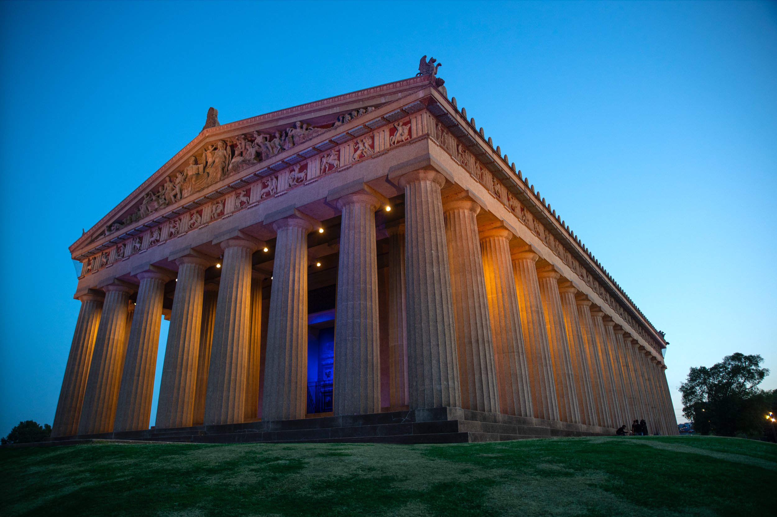 Nighttime view of the Parthenon, a historic Greek-style temple with tall columns, illuminated with warm lighting against a darkening sky, and surrounded by trees and a grassy area.