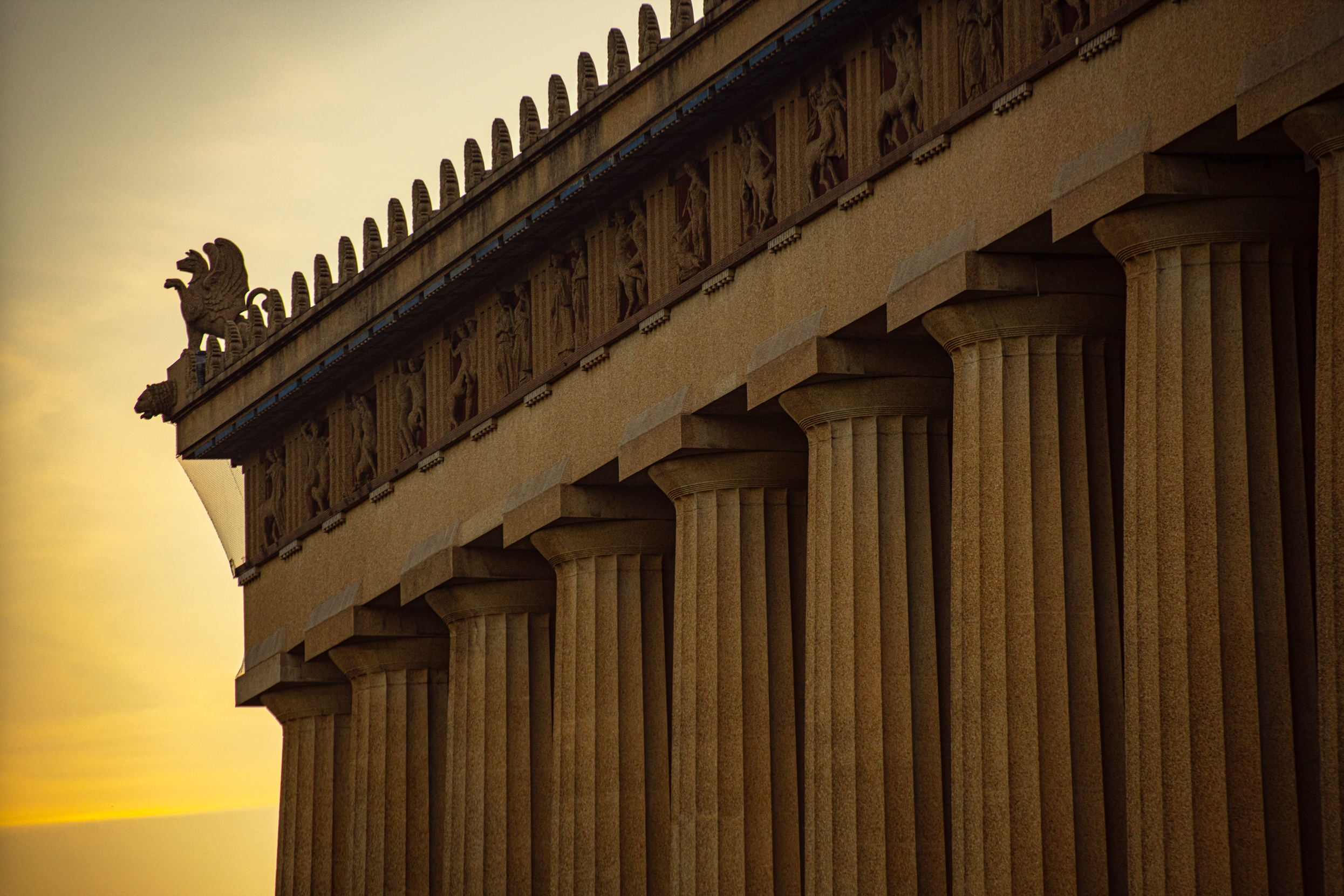 Close-up of the upper part of the Parthenon, a Greek historical temple, showing marble columns and detailed friezes, with a cloudy sky in the background.