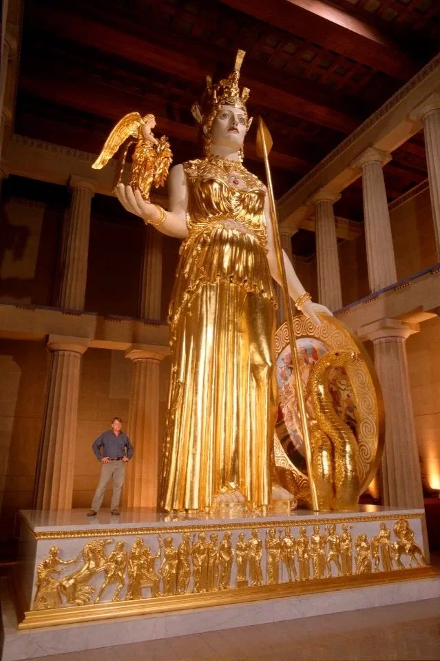 Large gold-colored statue of a woman in ancient Greek attire, holding a spear and a shield with a dragon design, surrounded by smaller gold statues on a decorative base, in a grand hall with tall columns and a wooden ceiling.  Athena Parthenos 