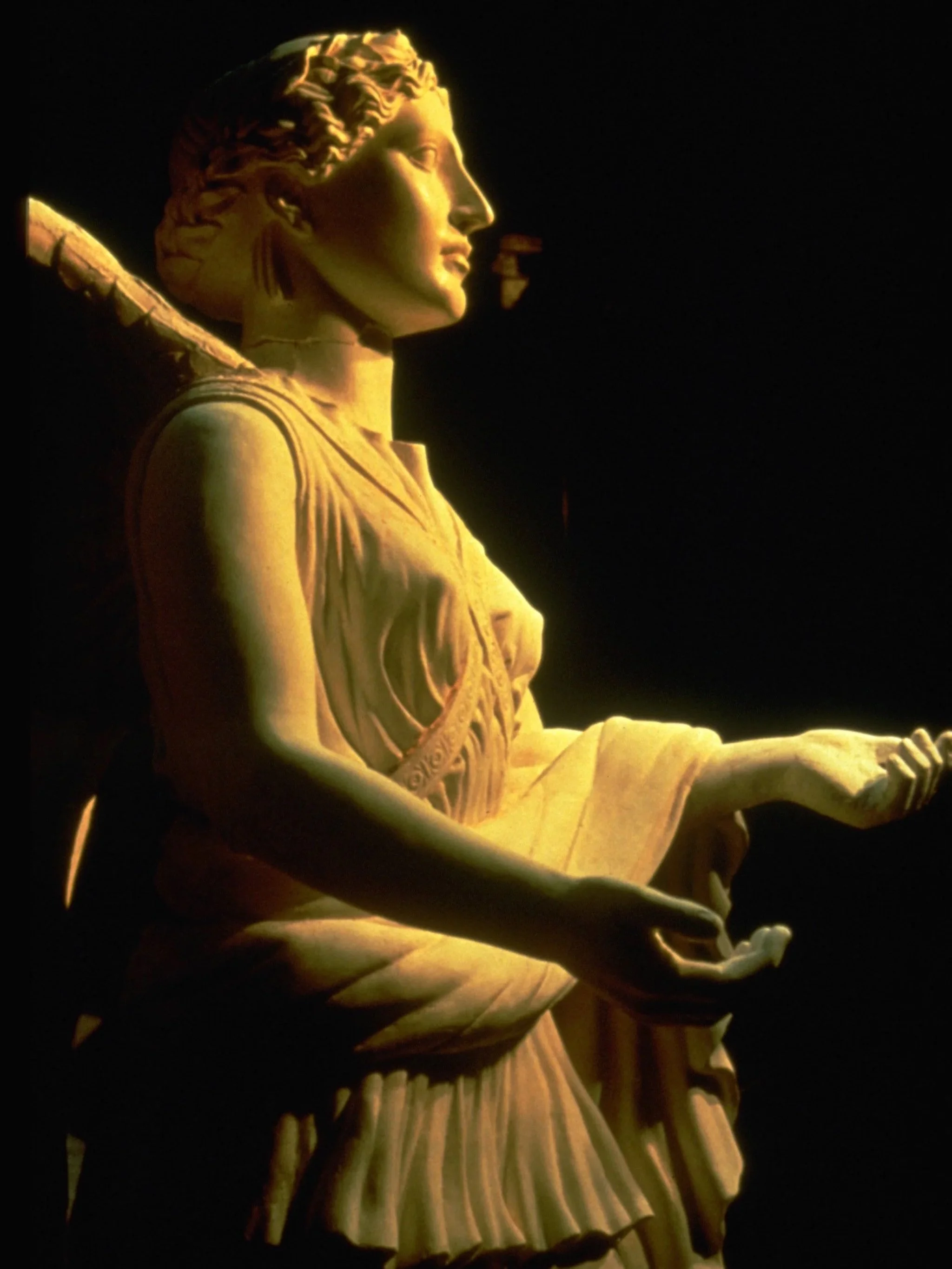 The illuminated statue of Nike at night, seen in profile with her head turned to the right, wearing a crown with rays and holding a tablet. Athena Parthenos in Parthenon Museum Nashville. 