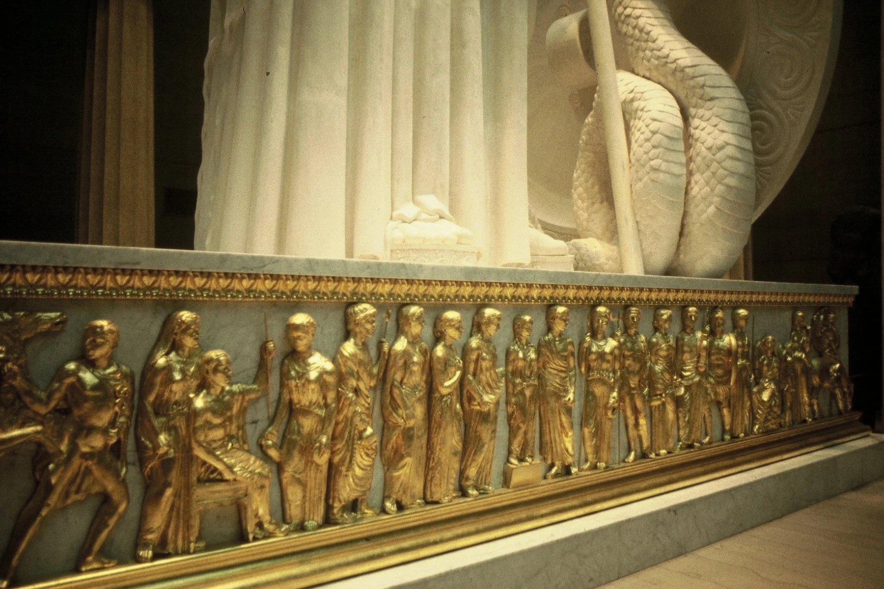 Golden sculpture depicting a procession of people and a large serpent sculpture in the background. Athena Parthenos in Parthenon Museum Nashville. 