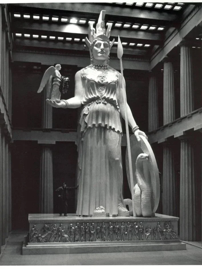 A large black and white sculpture of a goddess with multiple arms, holding a torch and a snake, with a smaller figure in her hand and a snake at her feet, located inside a building with columns and a wooden ceiling. Athena Parthenos in Parthenon 
