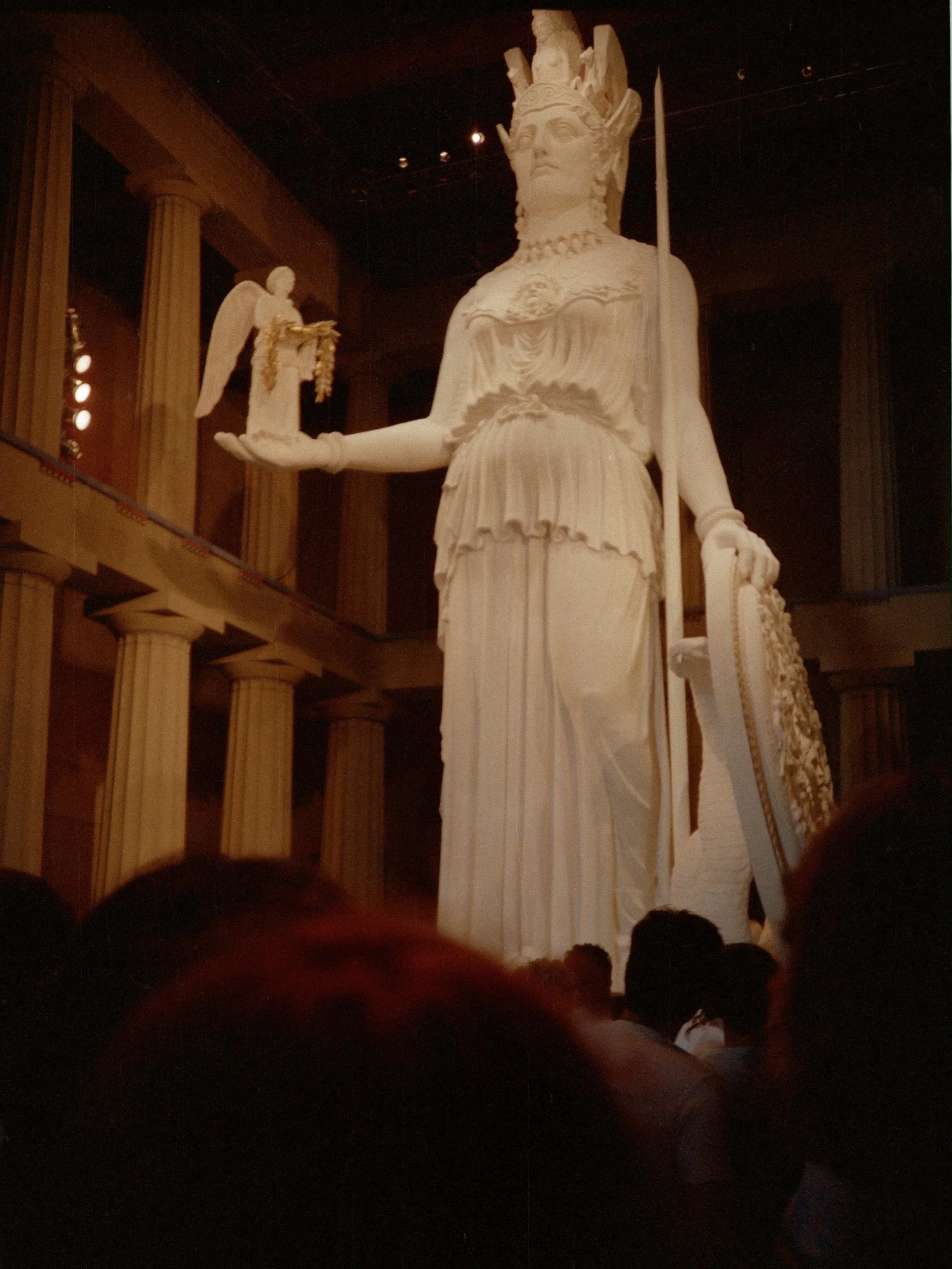 A large white statue of a goddess or warrior holding a sword in one hand and a smaller statue in her other hand, with a crown and elaborate clothing, displayed in a grand hall with tall columns. Athena Parthenos in Parthenon Museum Nashville. 