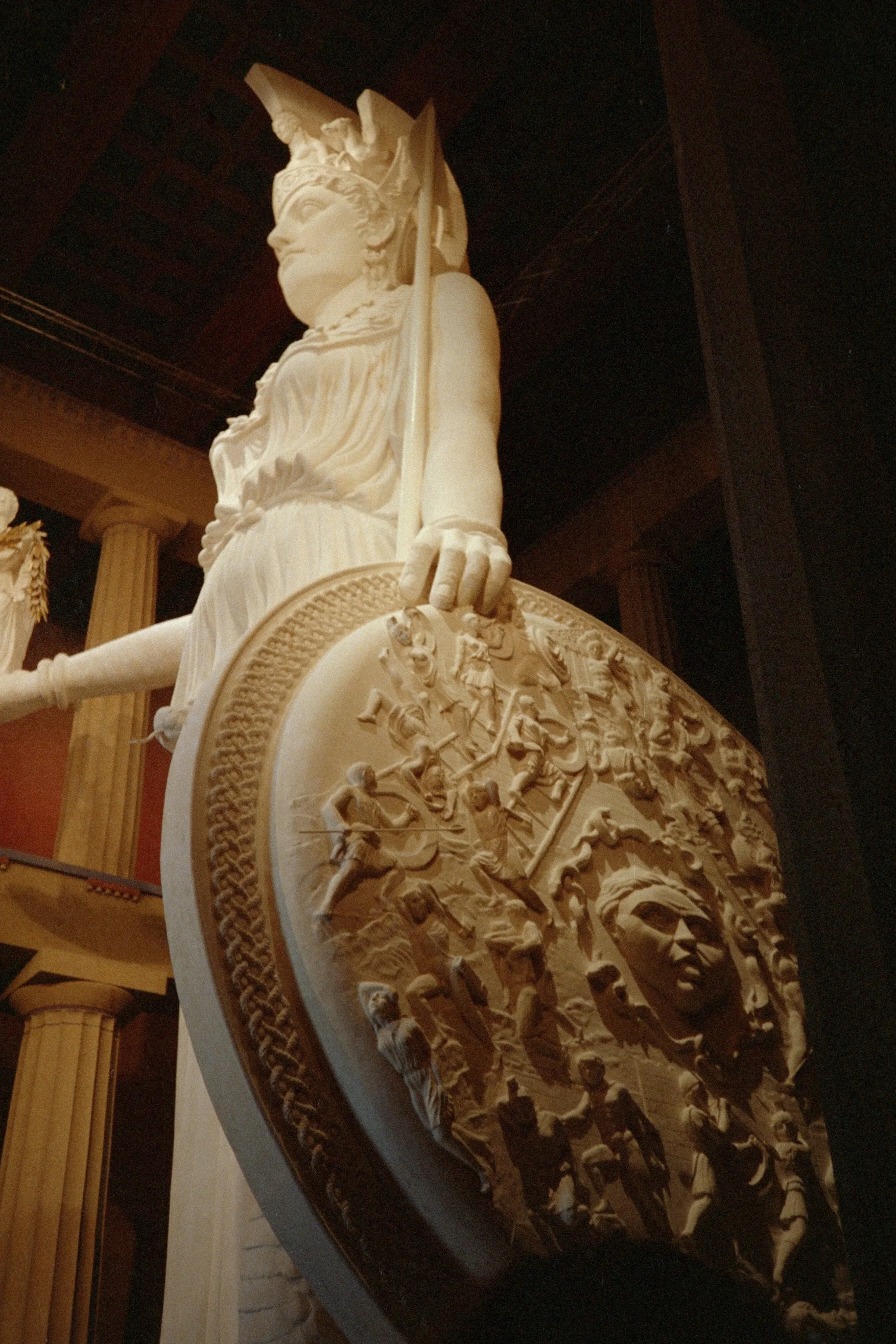 A large white sculptural frieze with detailed carvings of ancient figures and scenes, featuring a central figure with a helmet, on a curved shield or panel, against a dark background with wooden architectural elements. Athena Parthenos in Parthenon 