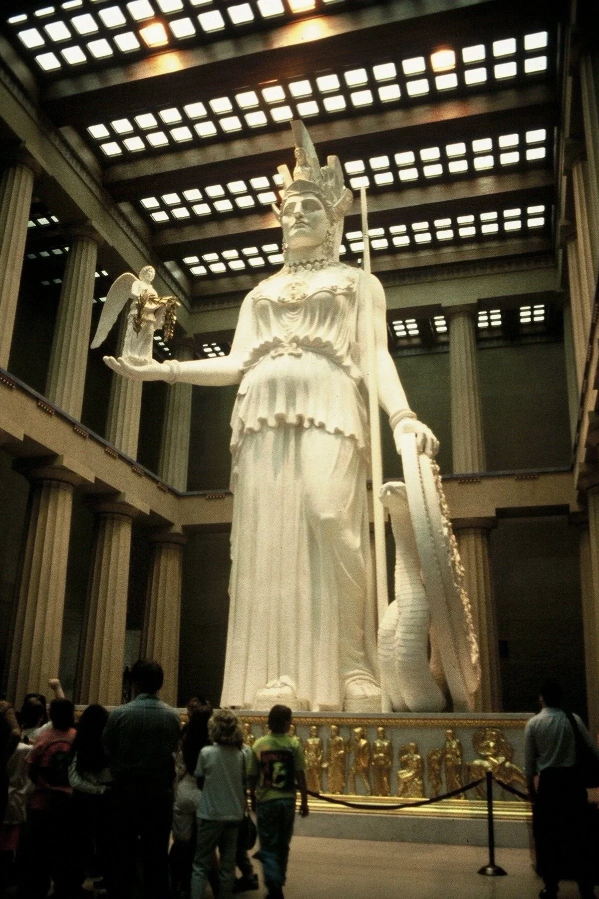 Large white statue of a woman with a crown and armor, holding a small angel statue in her right hand, standing inside a grand hall with tall columns and a ceiling with checkered light panels, surrounded by visitors. Athena Parthenos in Parthenon 