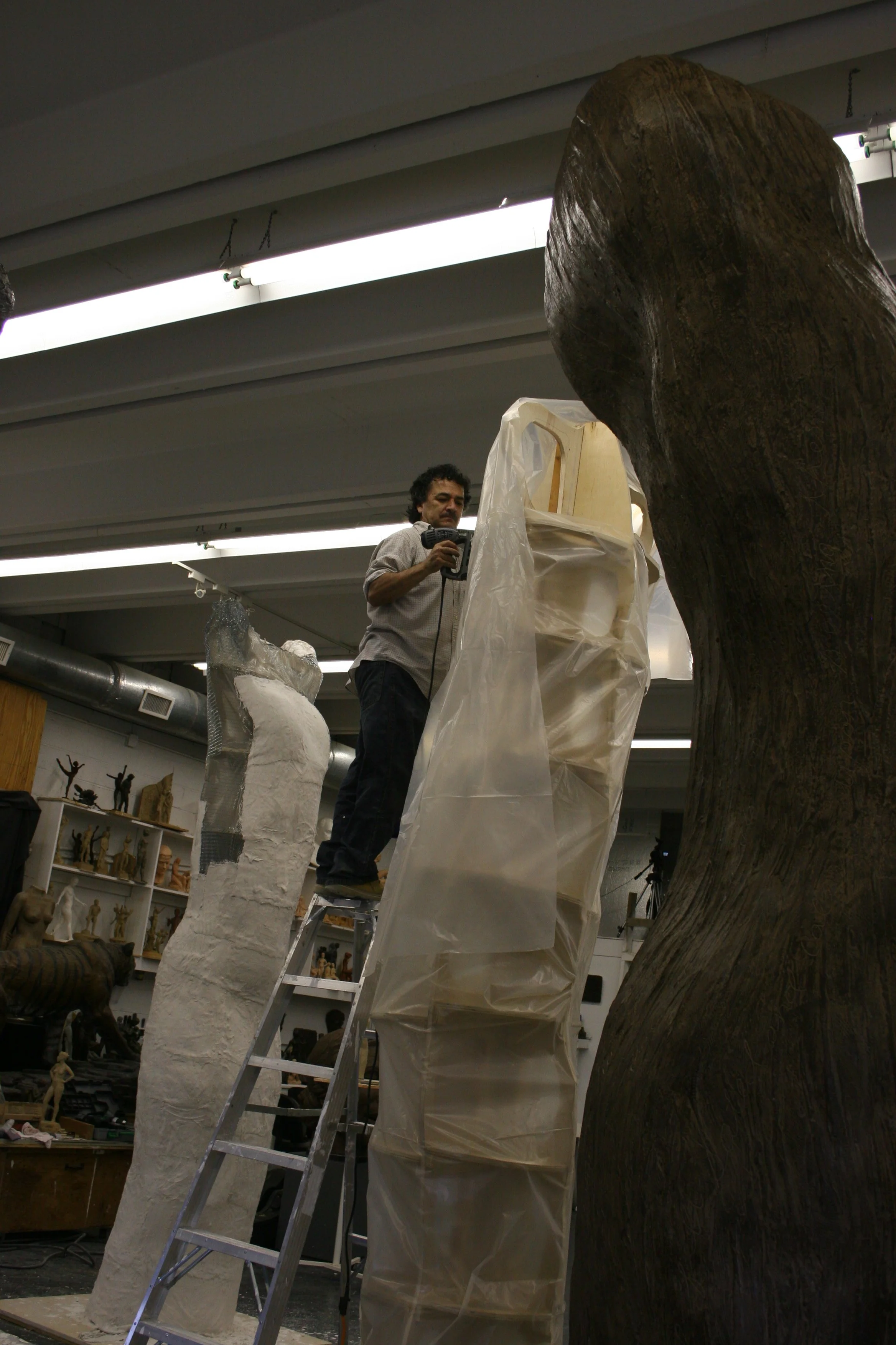 A man working on a sculpture in an art studio, standing on a ladder and using a power tool on a tall, white sculpture with metal covering. There are other sculptures and art supplies in the background.