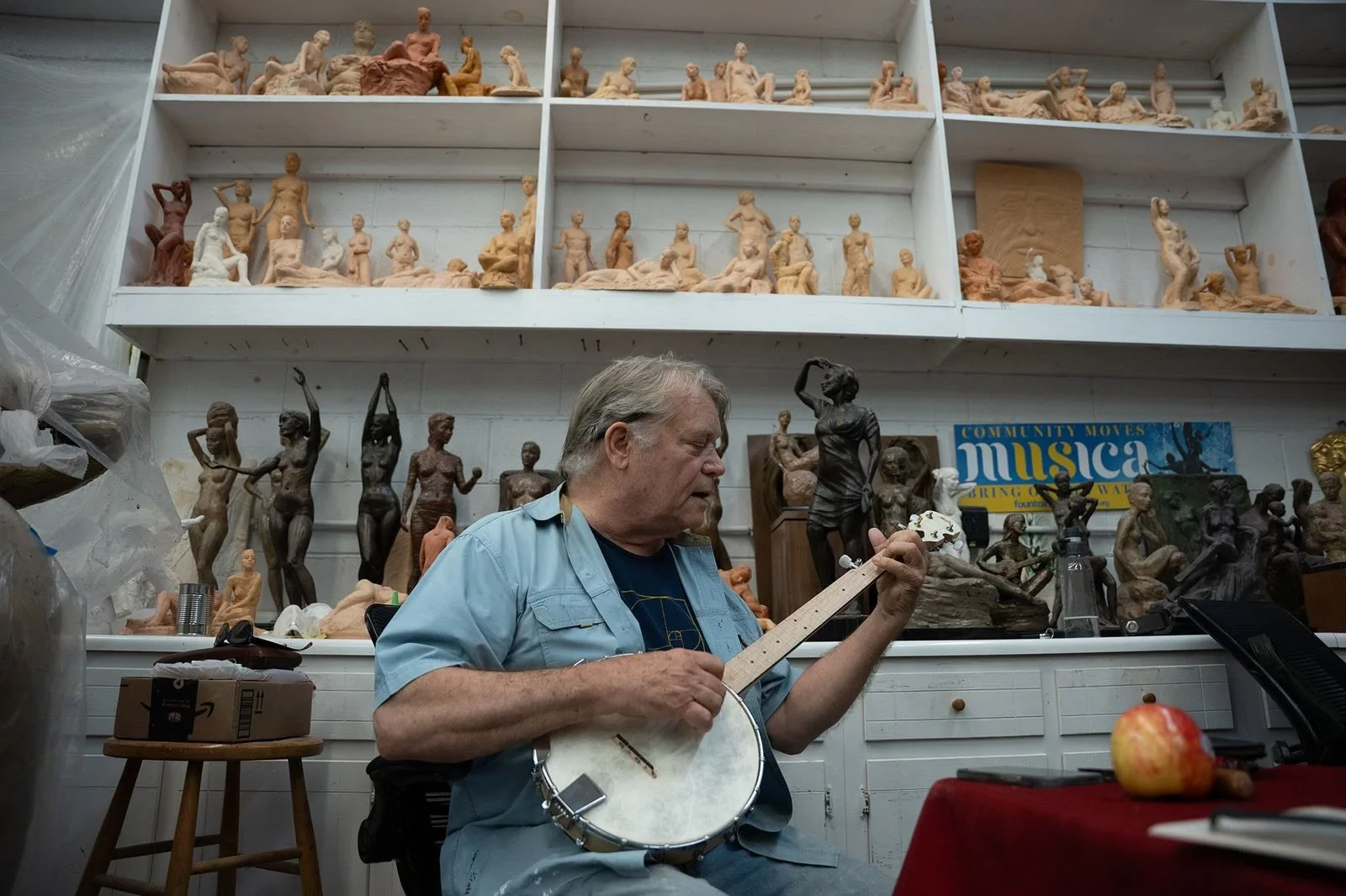 An older man playing a banjo in an art studio filled with sculpture models and artworks on shelves and walls.