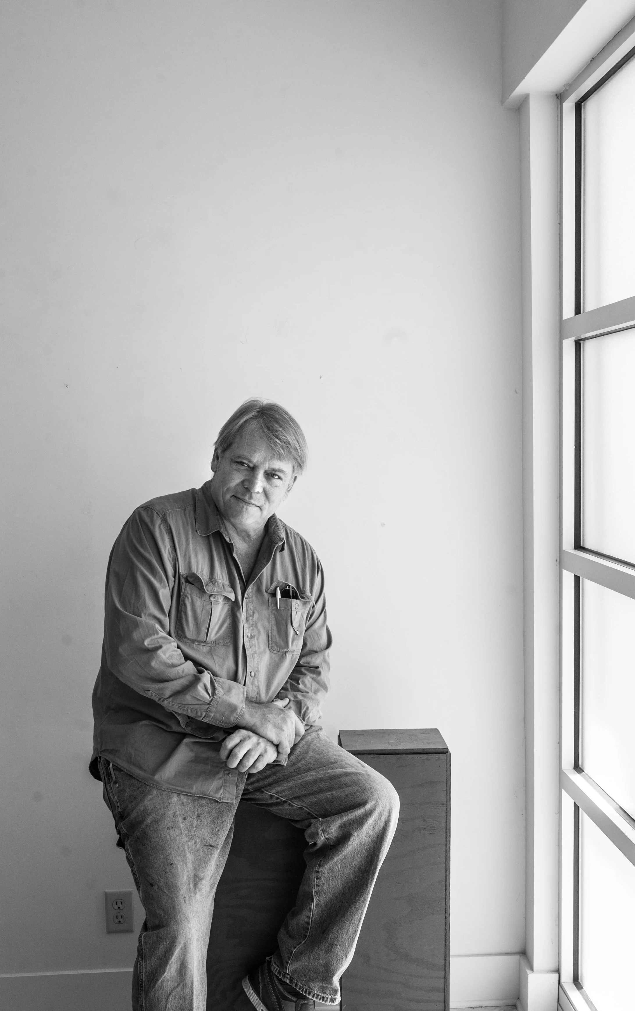 A man with light-colored hair and casual clothing sitting on a wooden box next to a window in a minimalist room.