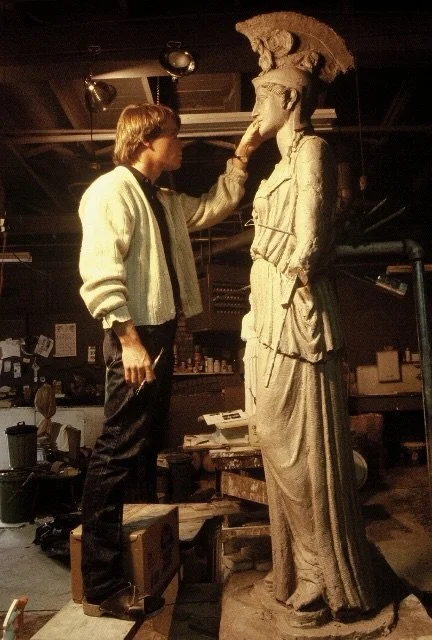 An artist or conservator examining an ancient Greek or Roman clay statue of a woman in a workshop or studio.