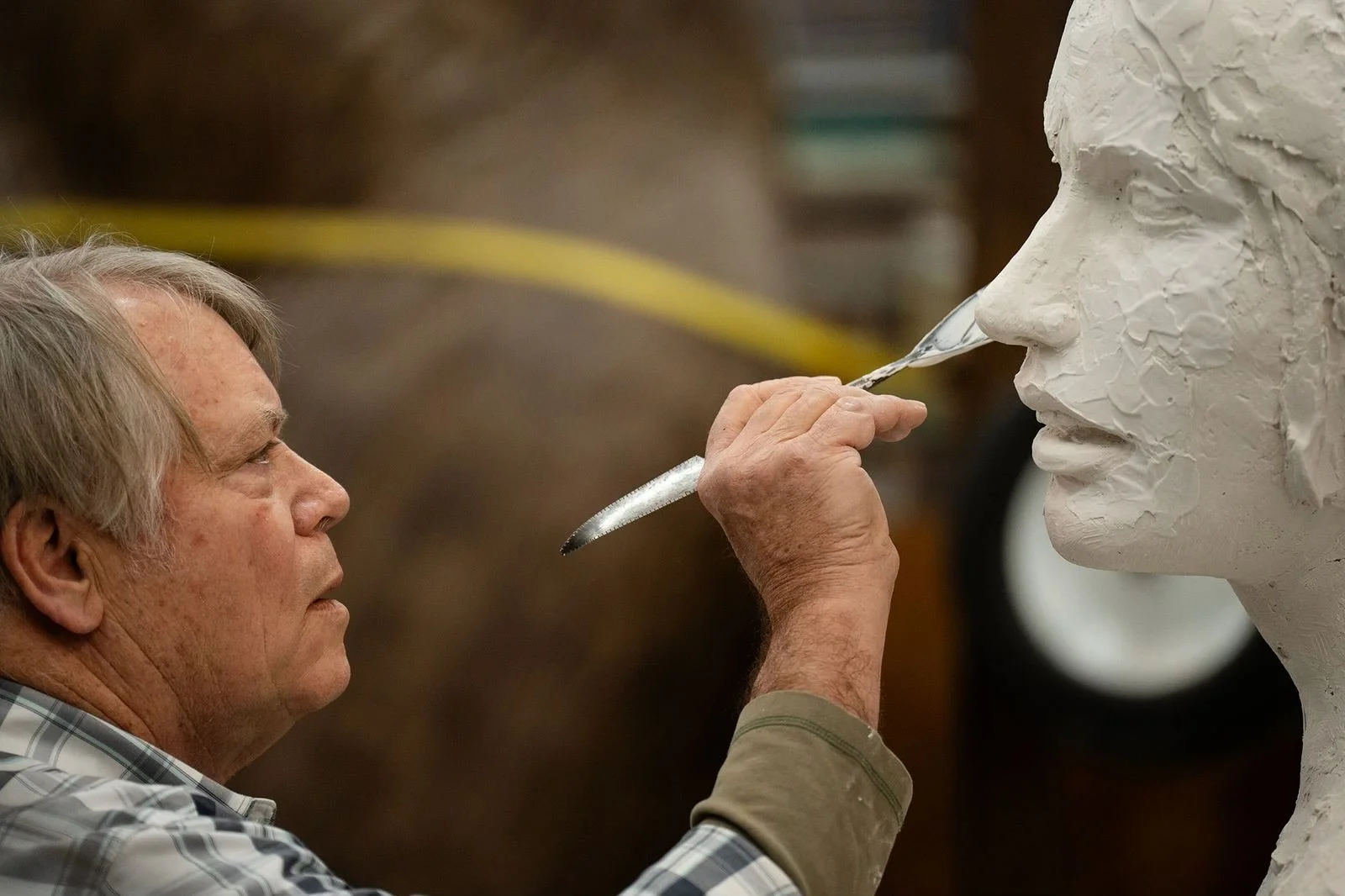 A man is sculpting a large, detailed bust of a person's face out of white material, possibly plaster or clay, in a workshop.