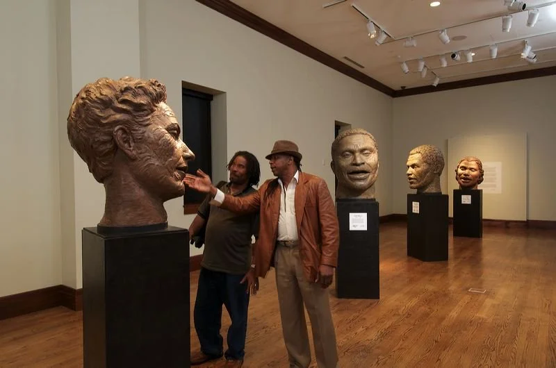 Two men are in an art gallery surrounded by bronze bust sculptures on black pedestals, with wooden floors and white walls.  Cultural Heroes