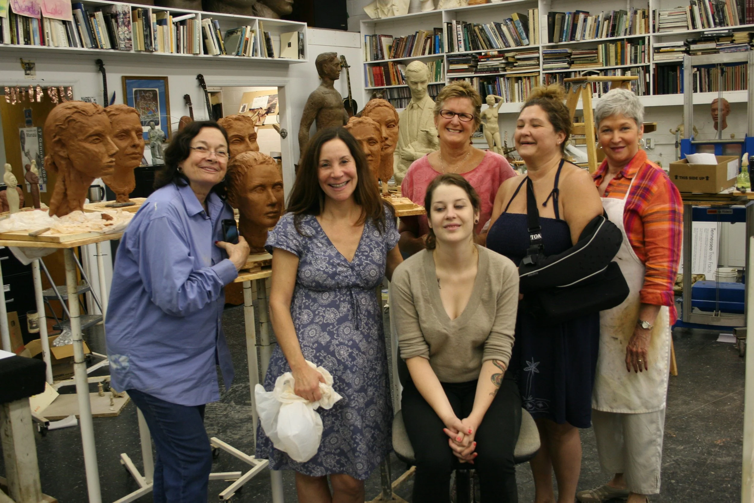 A group of women and a girl posing together in an artist's studio filled with sculptures, busts, and shelves of books.