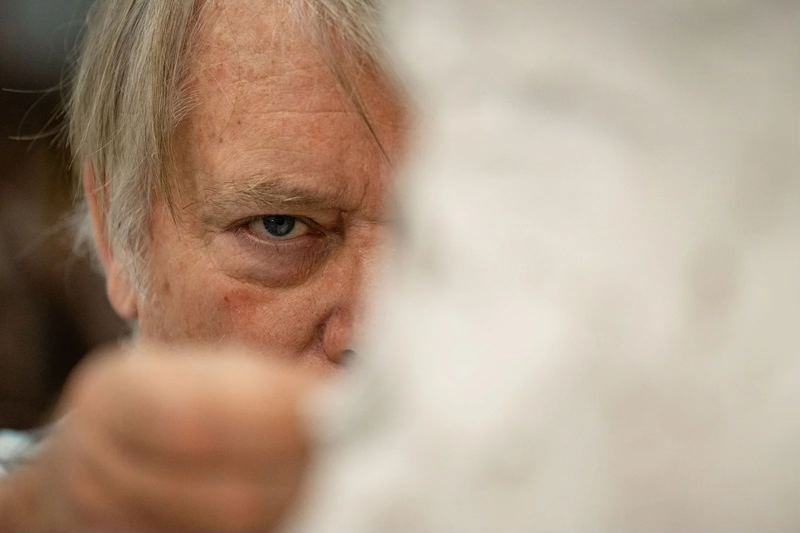 A close-up of a man with gray hair and blue eyes, partly obscured by a blurred object or hand in the foreground.