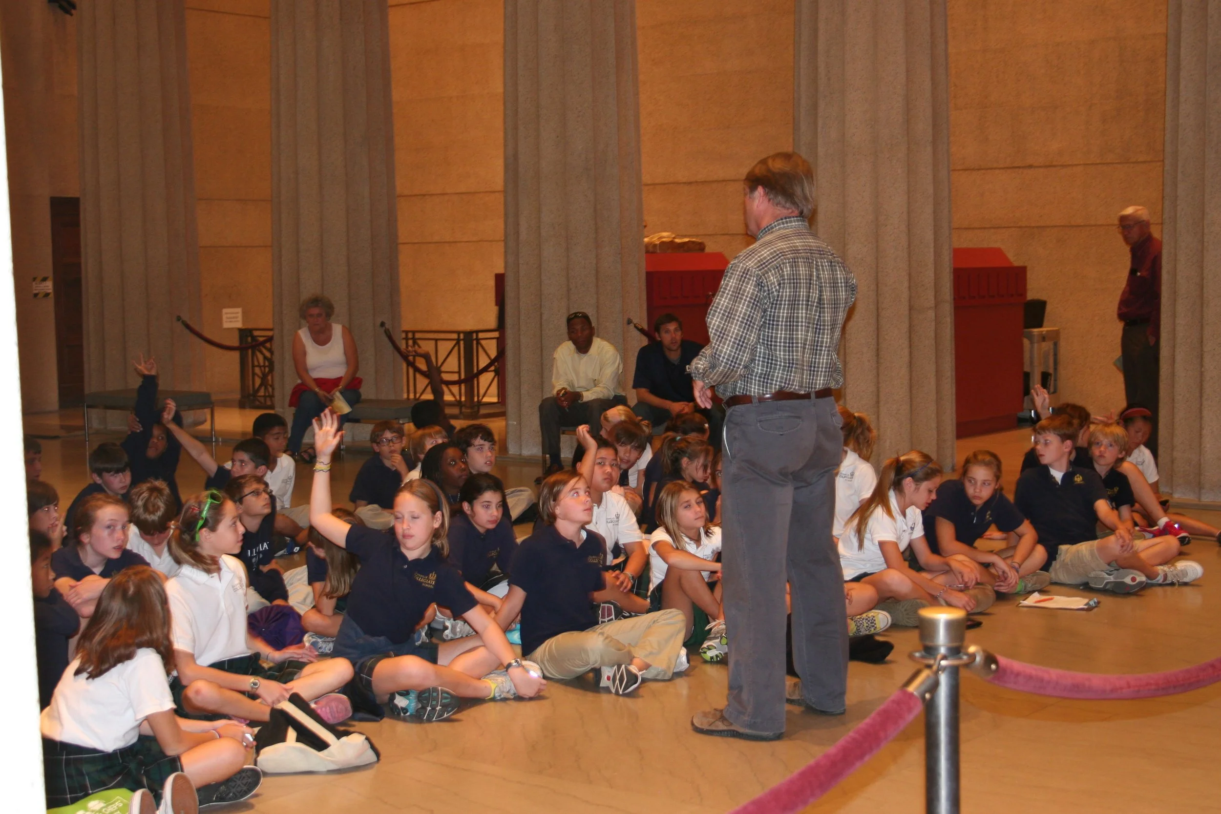 A man stands in front of a seated group of children in a large room with high ceilings and tall pillars. The children are sitting on the floor, attentively listening, with some raising their hands. Several adults are seated or standing in the backgro