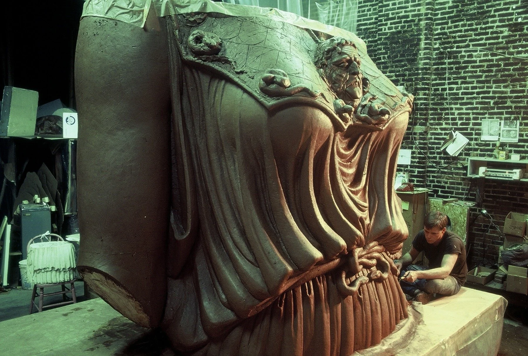 Two people are working on the sculpture in a dimly lit workshop with brick walls, shelves, and art supplies visible in the background. Athena Parthenos in Parthenon Museum Nashville. 