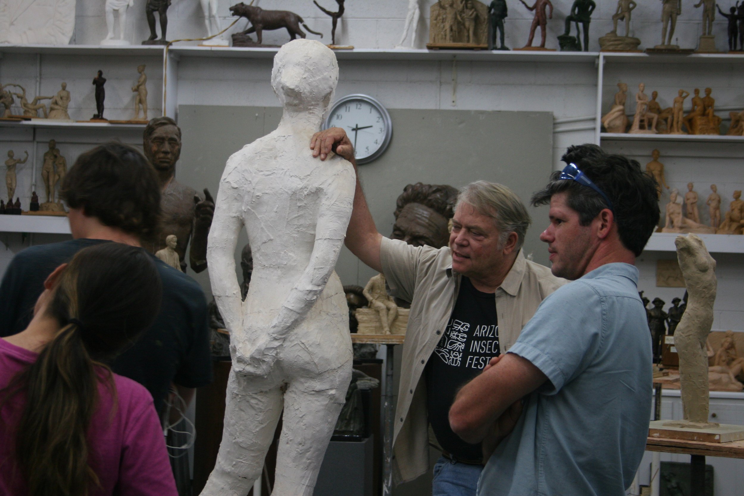 People working on a large white sculpture in an art studio surrounded by shelves filled with smaller sculptures and statues.