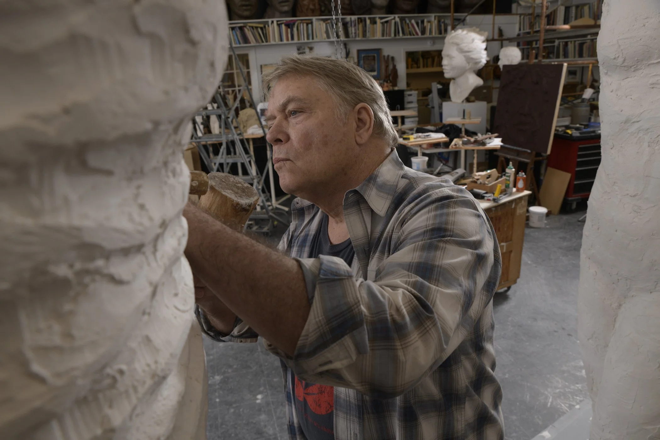 A man working in an art studio, shaping a sculpture between two white plaster walls, with bookshelves and bust sculptures visible in the background.