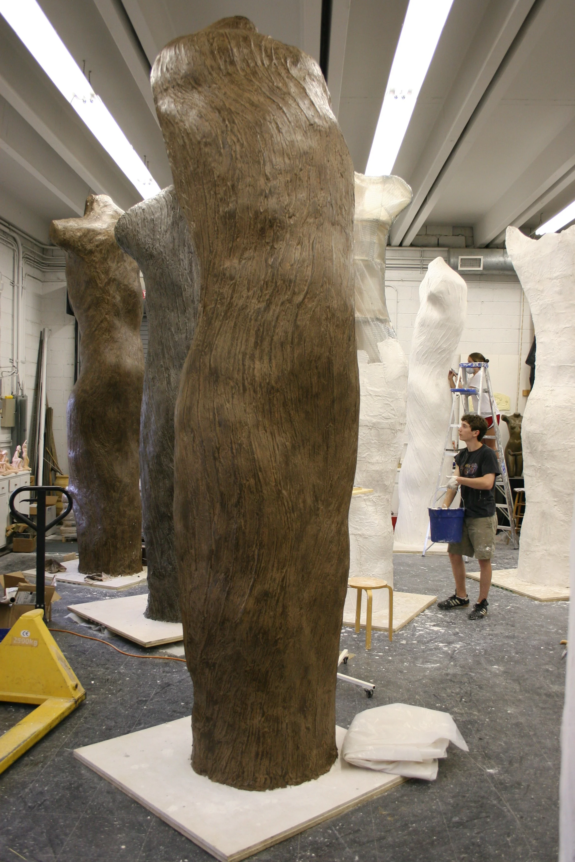 Individuals working on large plaster sculptures in an art studio or workshop. The sculptures resemble oversized tree trunks or abstract forms with natural wood textures. One person is on a ladder, and others are handling materials.