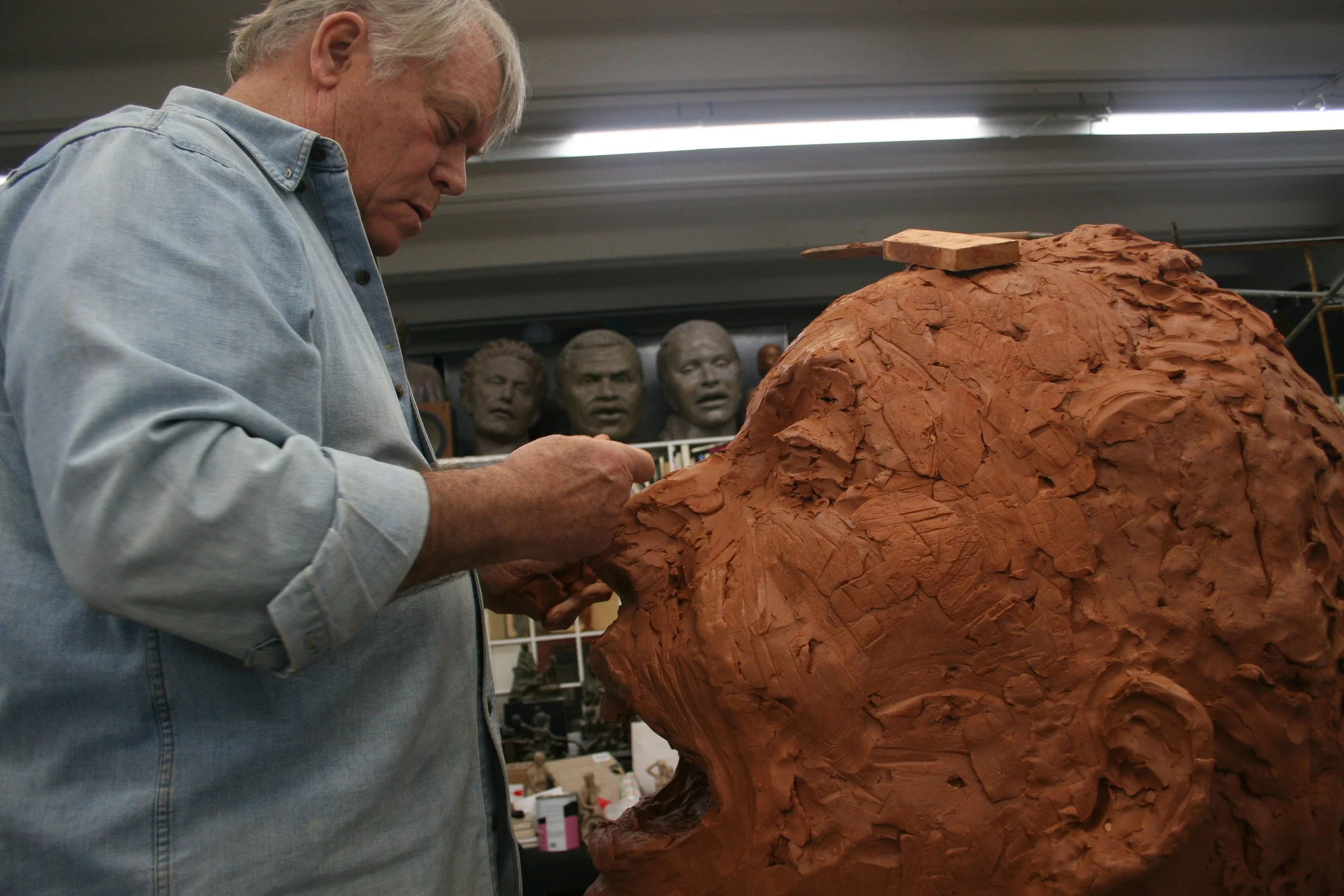 An artist working on a large clay sculpture Pete Seeger in a studio, with sculpted heads displayed in the background.
