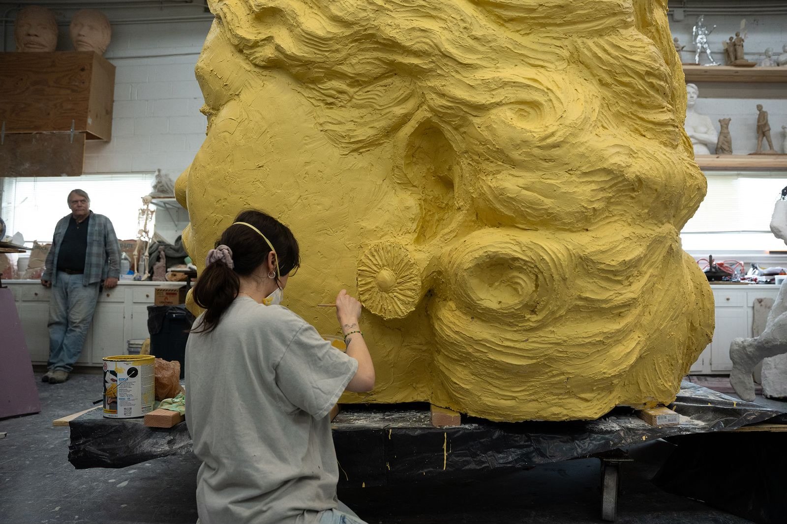 A person working on a large yellow sculpture of a face in an artist's studio, with a man standing in the background.