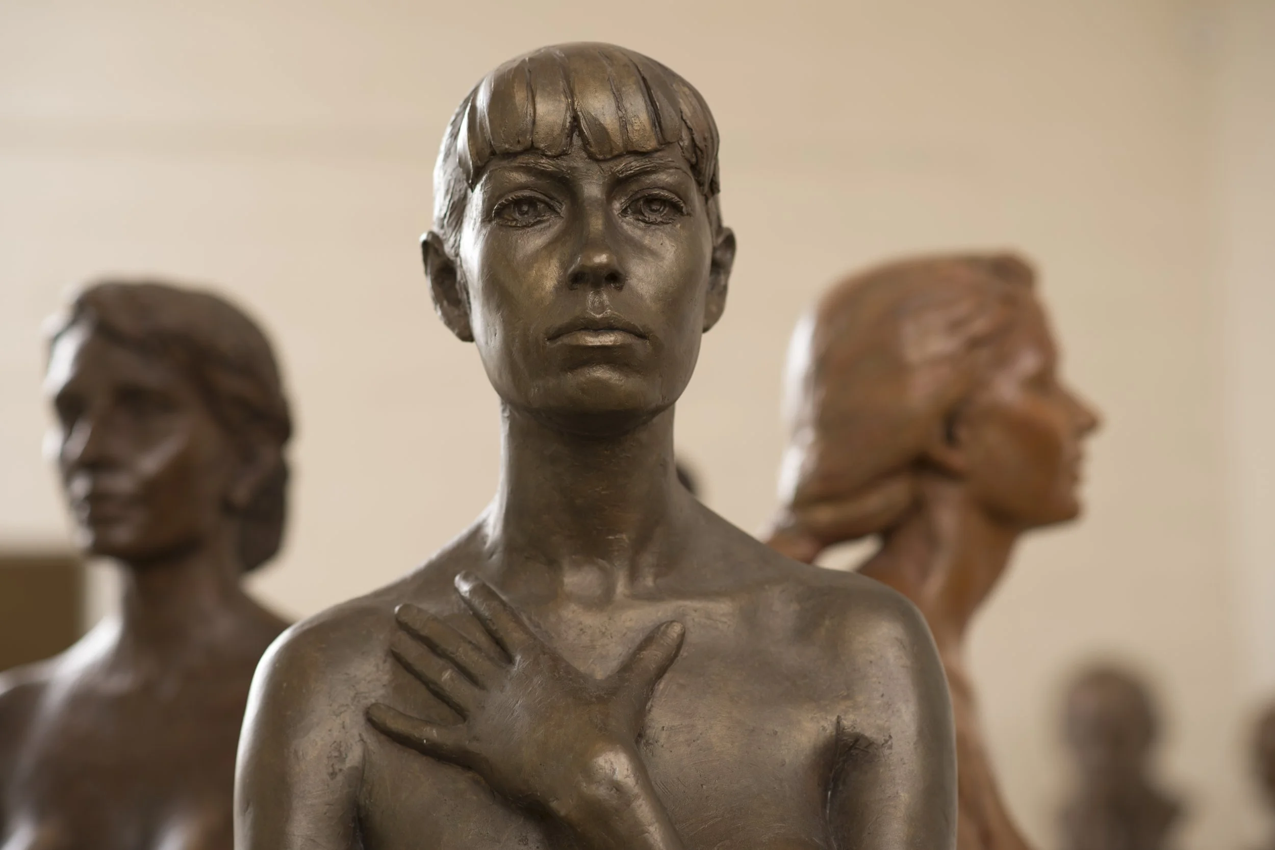 Close-up of bronze sculptures of three women with different hairstyles, shoulders, and hands crossed over their chests, on display in a gallery.