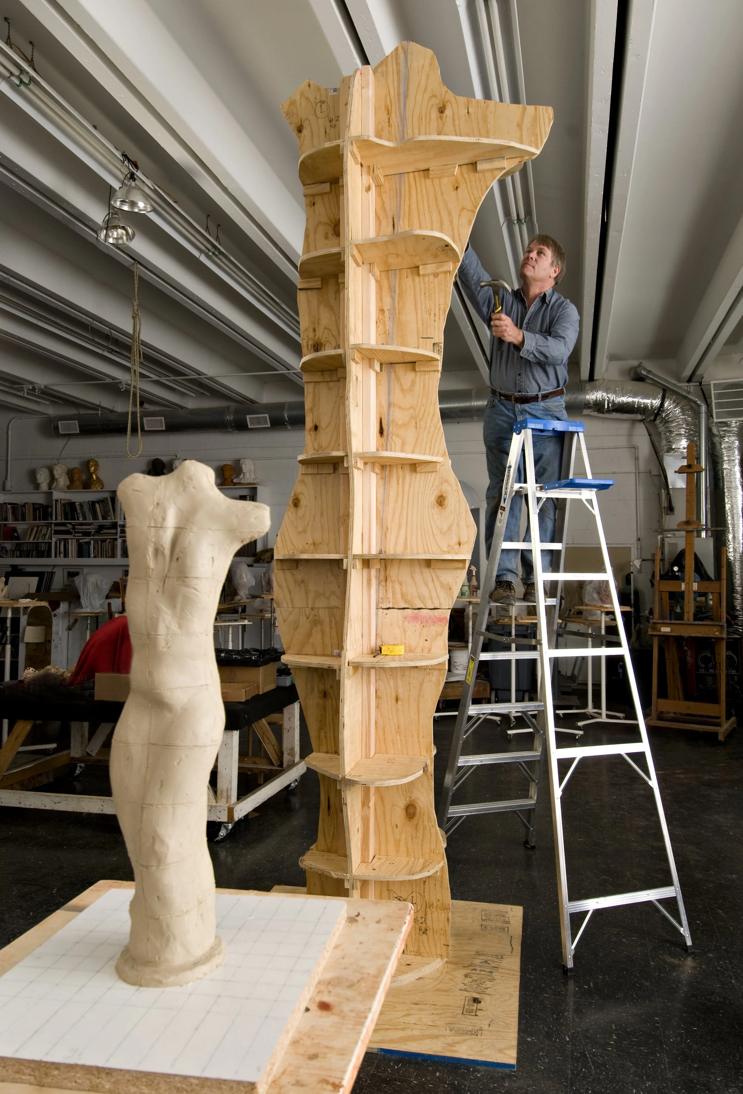 A man works on a wooden sculpture in a studio, with shelves of books and art busts in the background. The sculpture is tall and narrow with a curving, abstract shape.