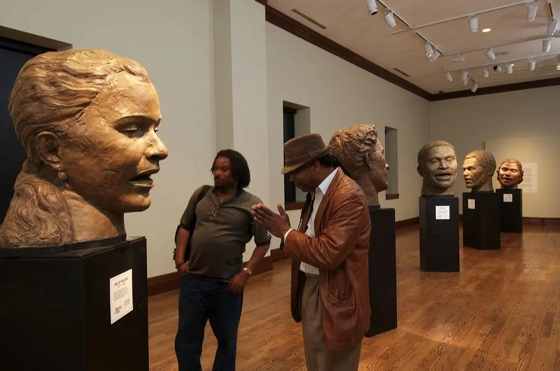 Two men observing and discussing bronze busts of notable African American figures displayed in an art gallery with wooden floors and white walls.  Cultural Heroes