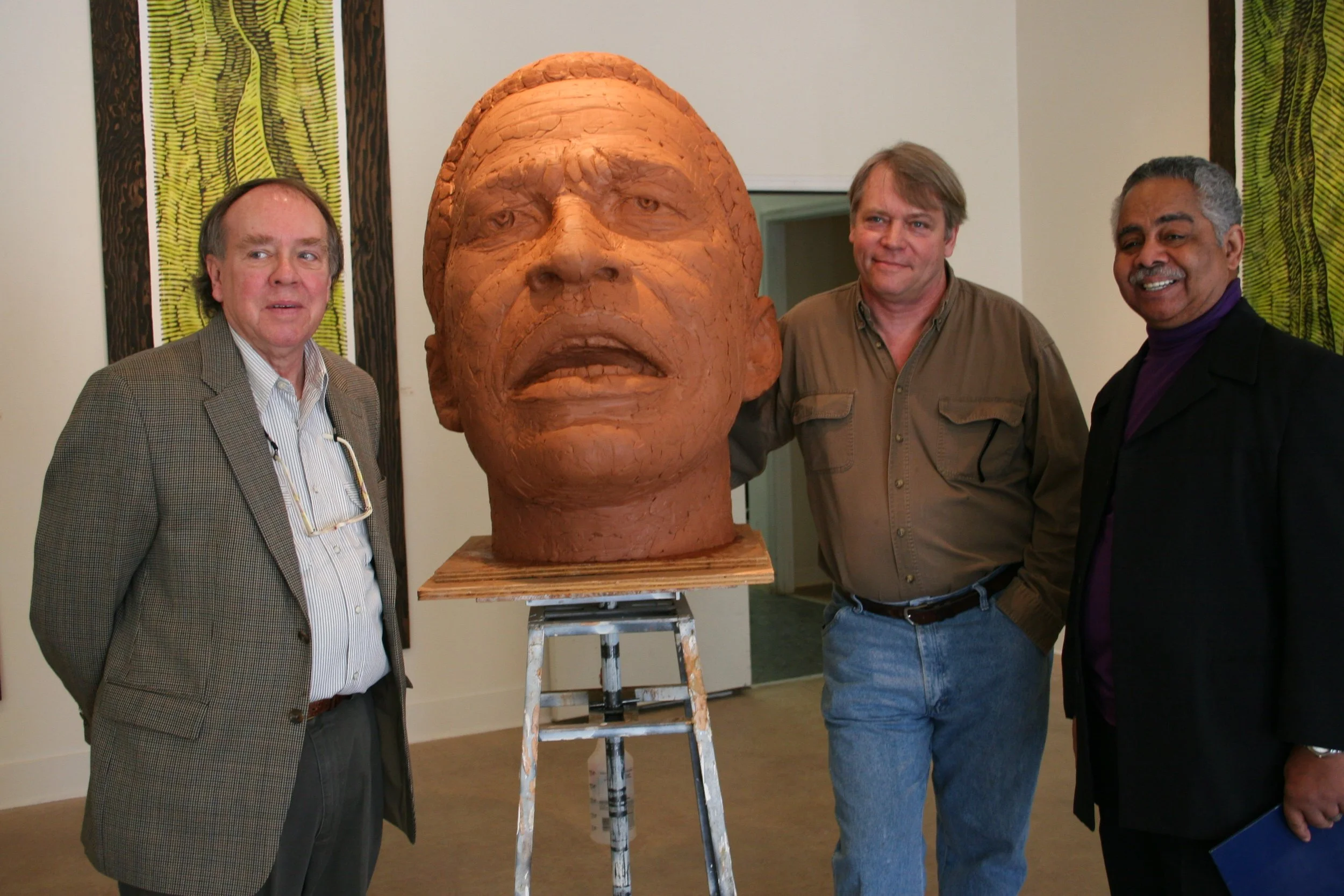 Four men standing around a large, realistic head sculpture made of a clay-like material in an art gallery. The sculpture has detailed facial features and appears to be of an older person. The men are smiling and posing for the photo.
