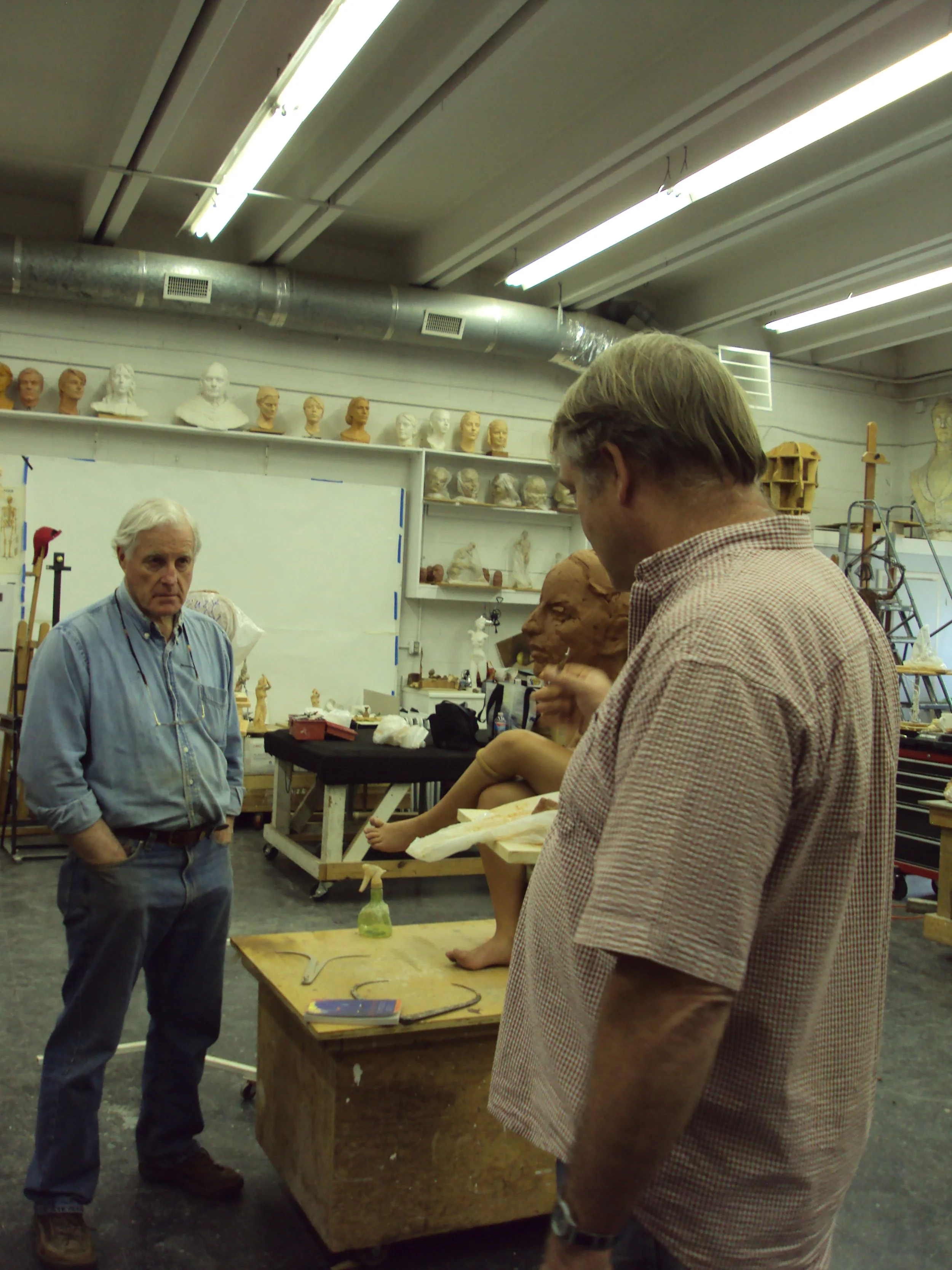 Two men in a workshop or studio, one with white hair and a blue shirt, the other with brown hair and a checkered shirt, stand near a wooden work table with sculptures and art supplies. The workspace has shelves with busts and bust sculptures, and var