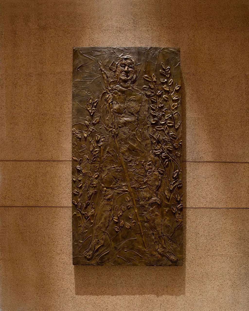 Bronze sculpture of a smiling woman holding a fishing pole, surrounded by detailed plant and foliage engravings, mounted on a wall.
