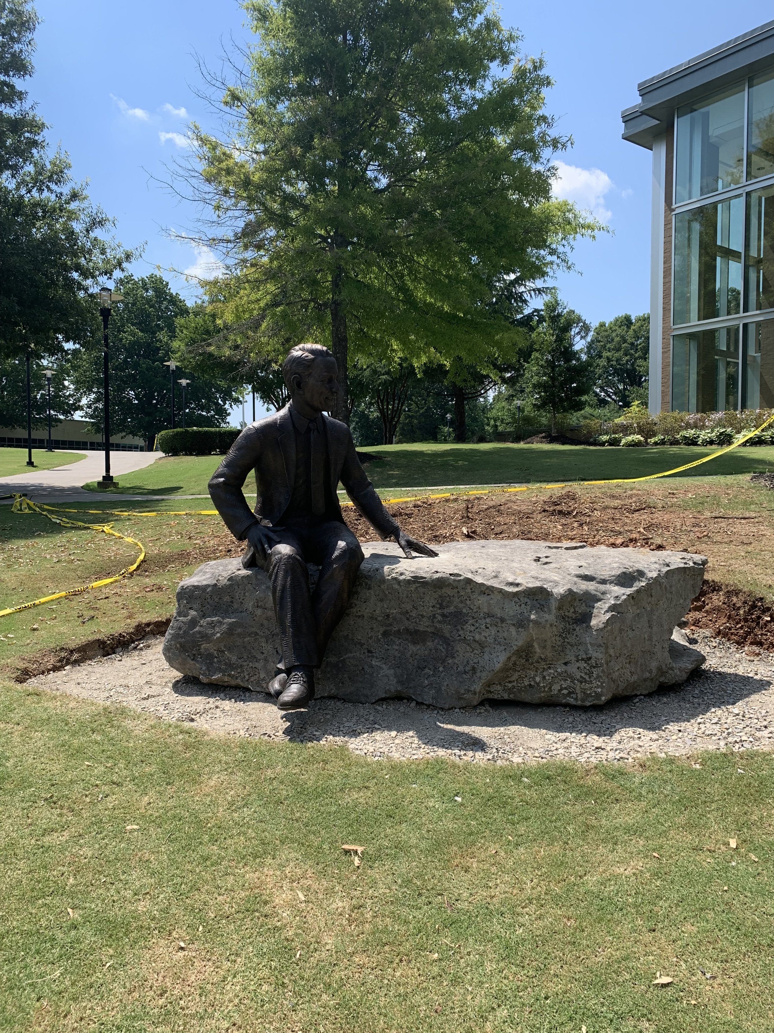 Statue of a seated man in a suit, placed on a large rock outdoors, with green lawn, trees, a modern glass building, and a blue sky in the background.