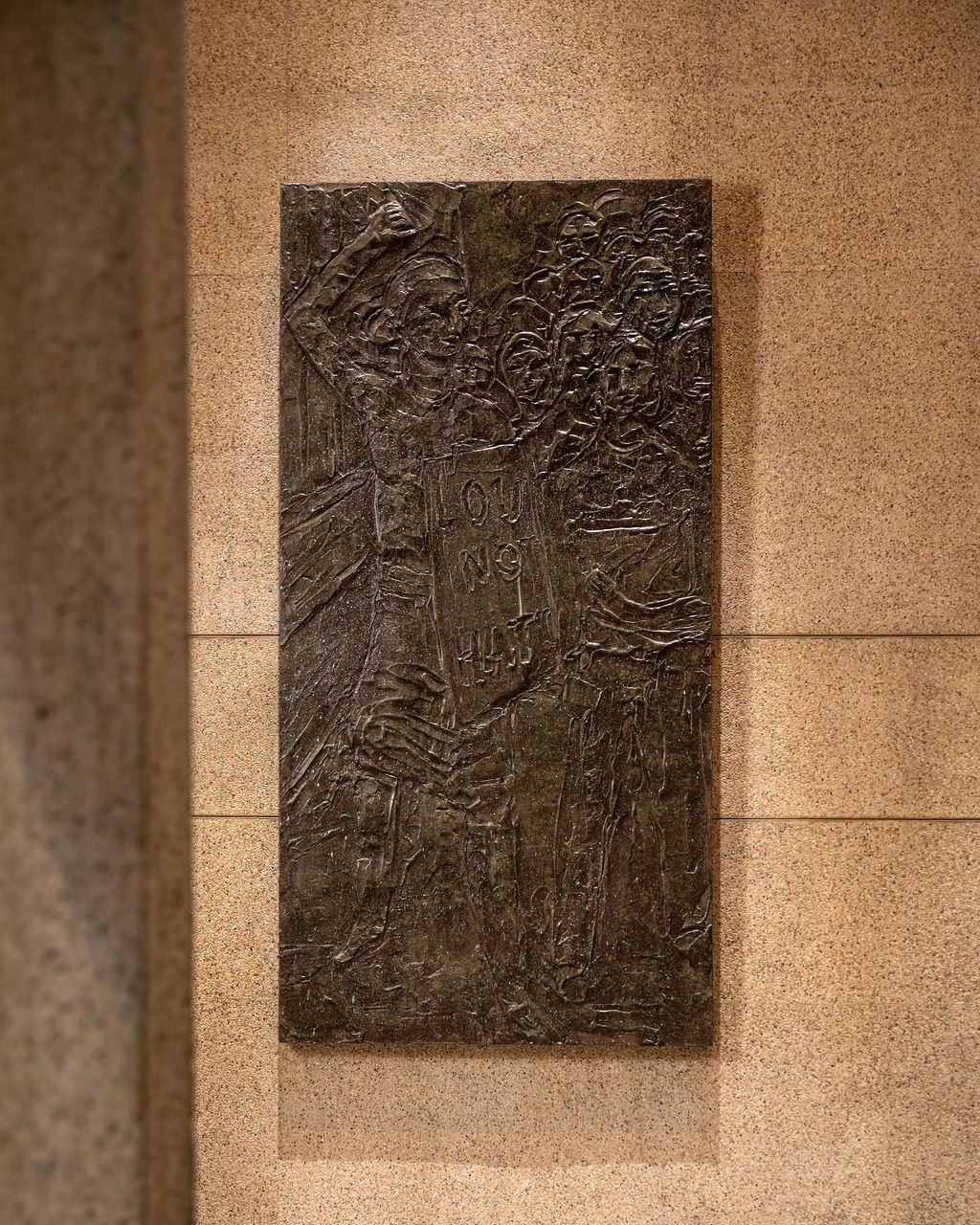 Bronze relief sculpture of female protesters, one holding a sign, on a beige wall.