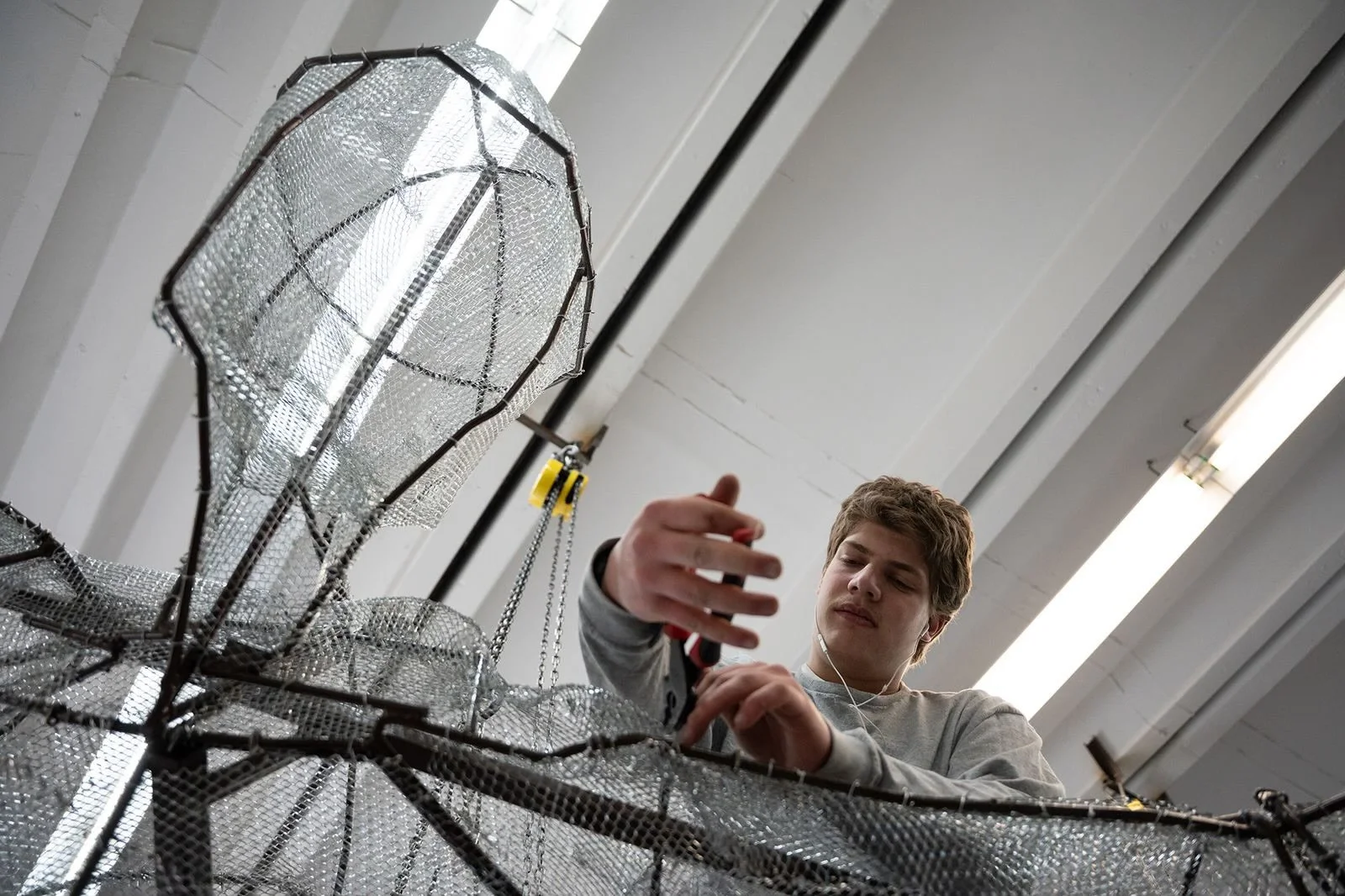 A young man working on a large metal and mesh structure in a well-lit industrial workspace.