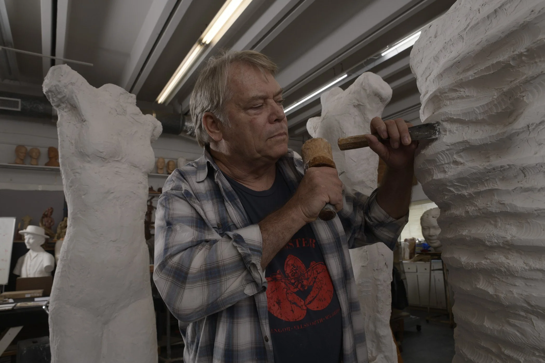 An elderly man working on a sculpture in an art studio, carving details into a large white plaster sculpture using a chisel and hammer.