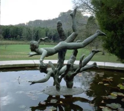 A sculpture of a girl running and jumping in a natural outdoor setting with trees and a pond.