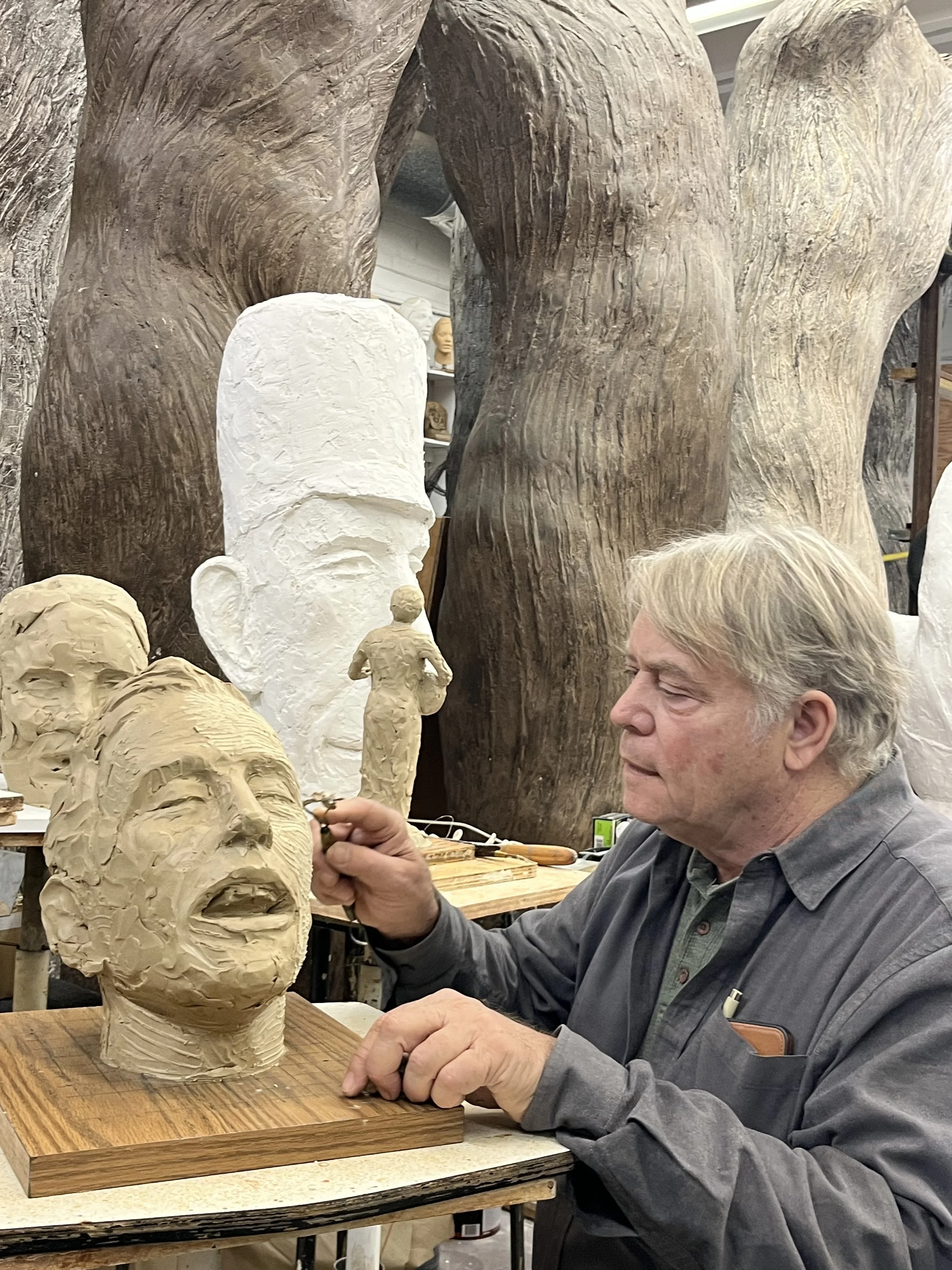 An artist sculpting a large clay head in a studio, with smaller sculptures and busts in the background.