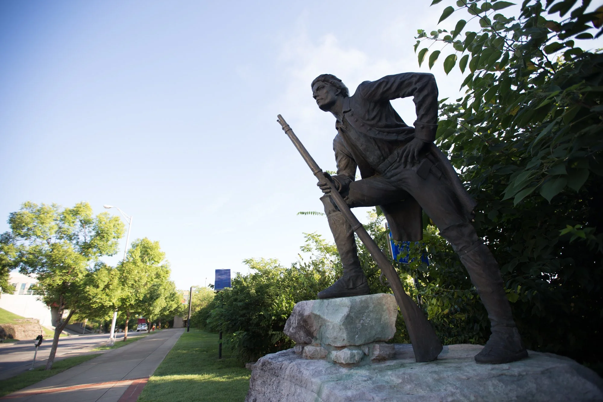 Bronze statue of a man in historical clothing, holding a rifle, standing on a stone base in a park with trees and a sidewalk in the background during daylight.