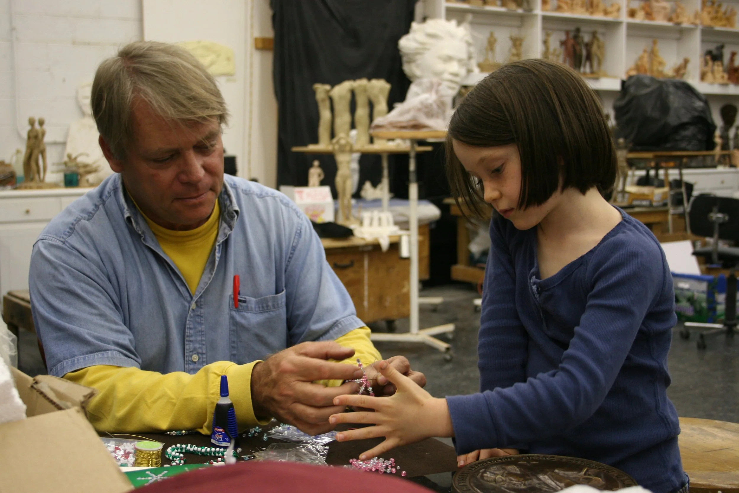 A man and a young girl working together on craft projects at a table in an art studio. The man is wearing a denim shirt and the girl is wearing a blue top. There are various craft materials like beads, glue, and jewelry on the table.