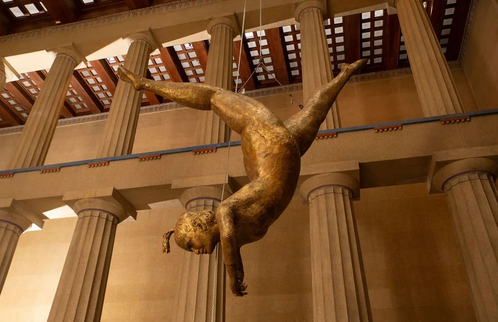 A gold-colored sculpture of a young girl hanging upside down in a museum with classical columns and a wooden ceiling.