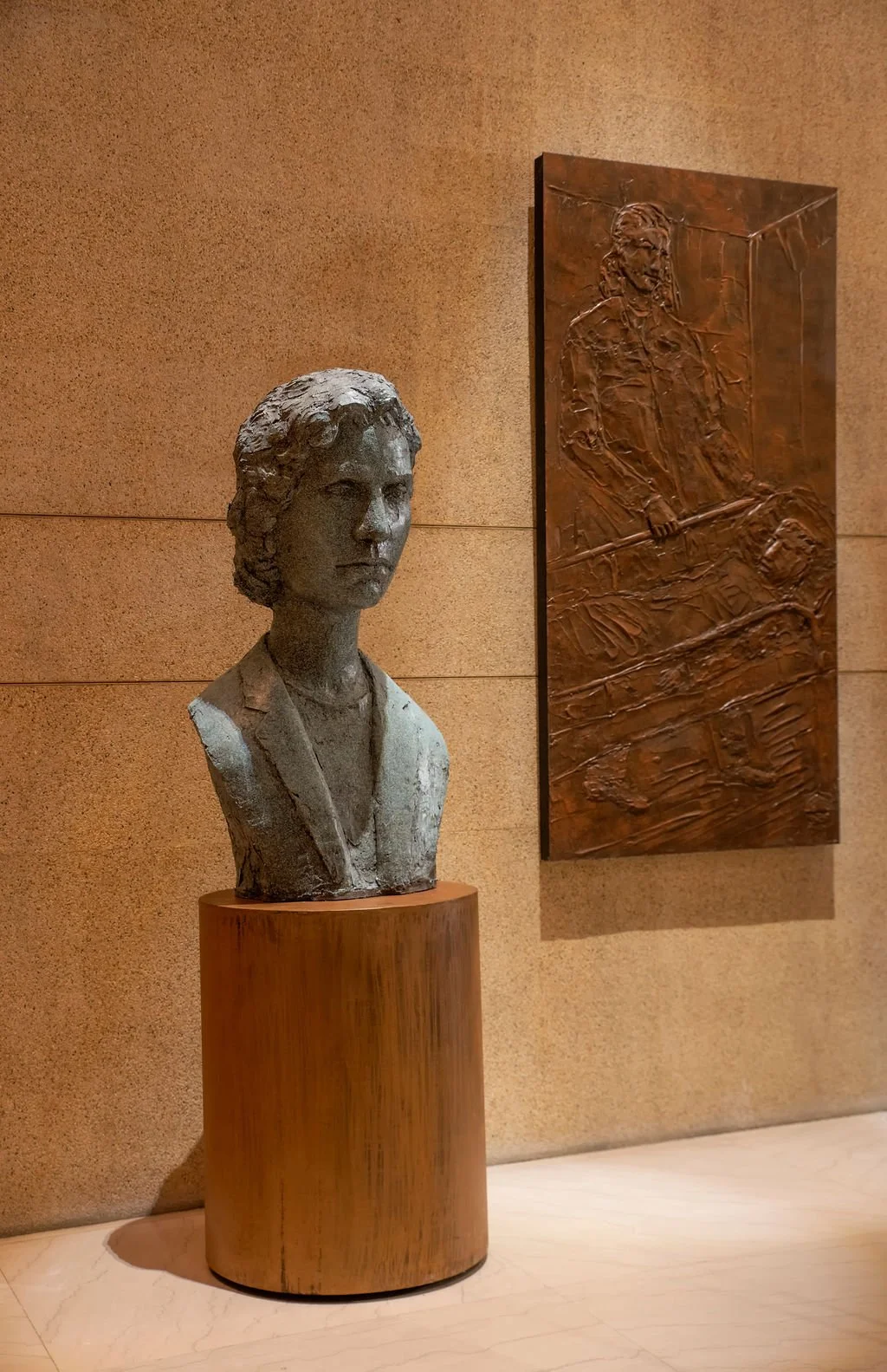 A plaster bust of a woman on a metal pedestal next to a bronze relief sculpture of a woman holding a hammer, displayed on a beige wall in an indoor setting.
