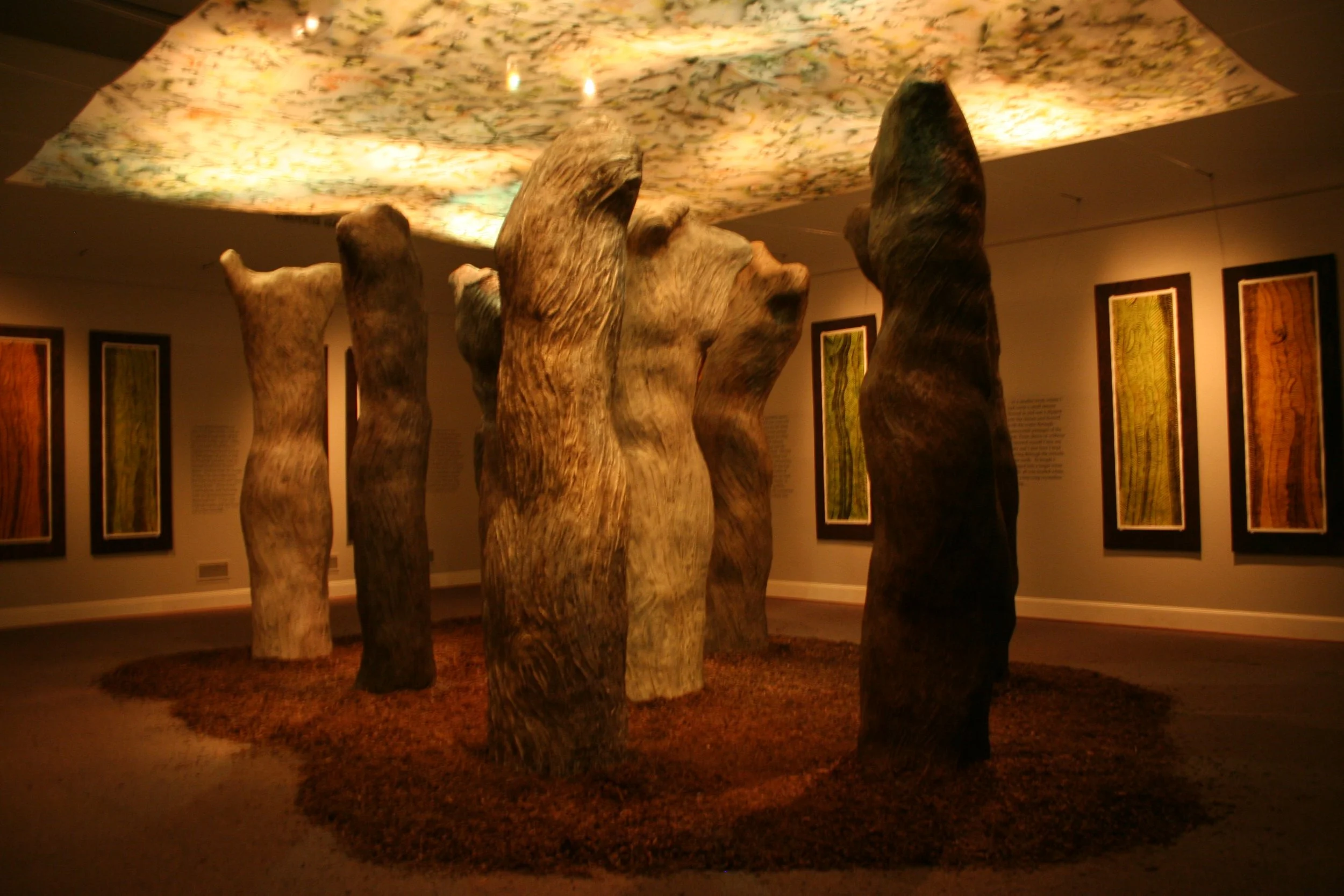 An art installation featuring several large, tree trunk-like wooden sculptures arranged in a circle on a carpeted floor, with framed artwork hanging on the walls in the background. Dream Forest Installation.