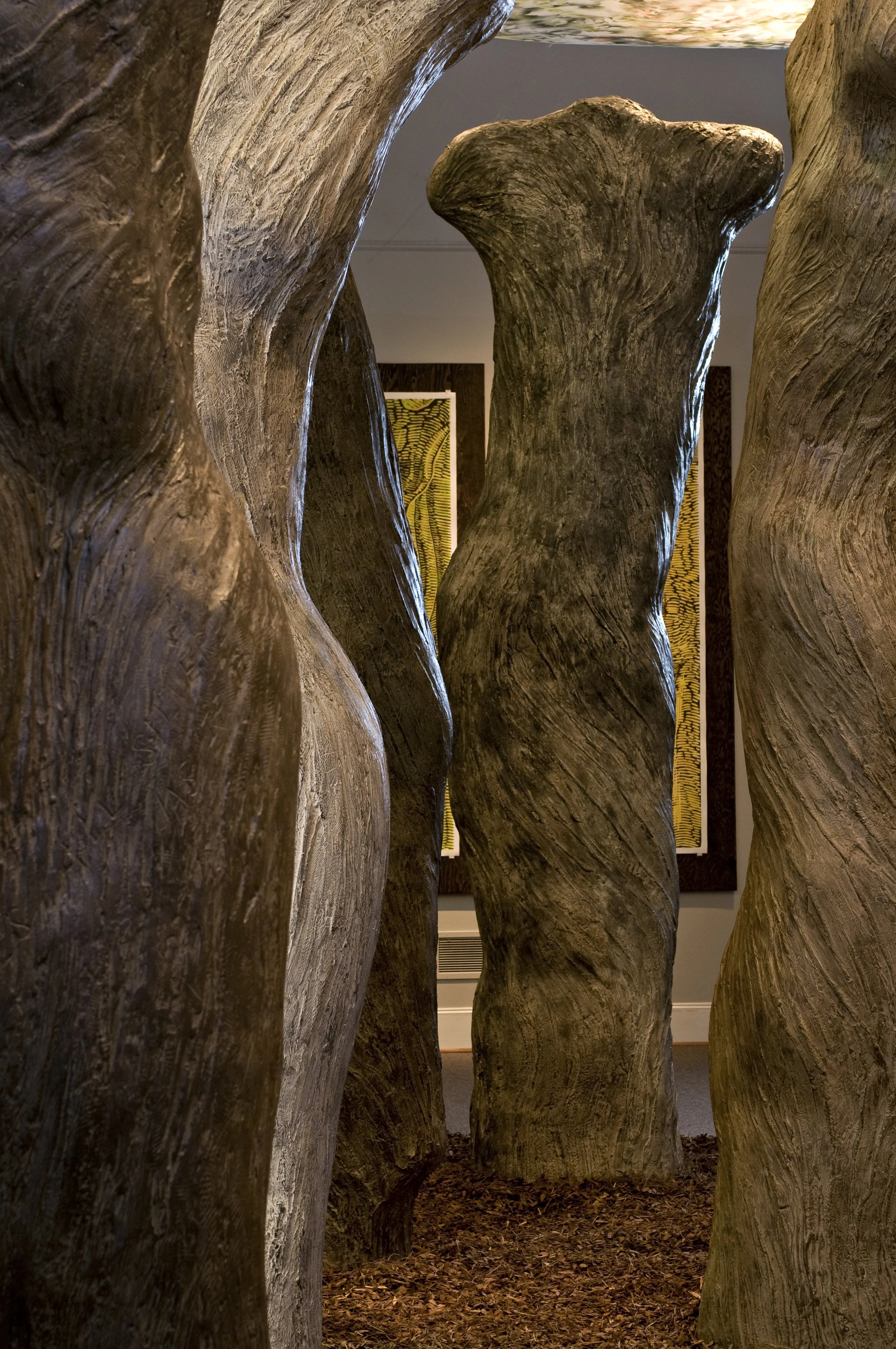 Close-up view of large, textured, wooden tree trunks arranged vertically, creating a narrow passage. The ground is covered with brown fallen leaves, and in the background, framed artwork is partially visible on a wall.