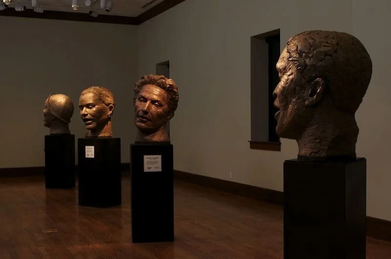 Four bronze bust sculptures of human heads on black pedestals inside an art gallery with wood floors and white walls.  Cultural Heroes