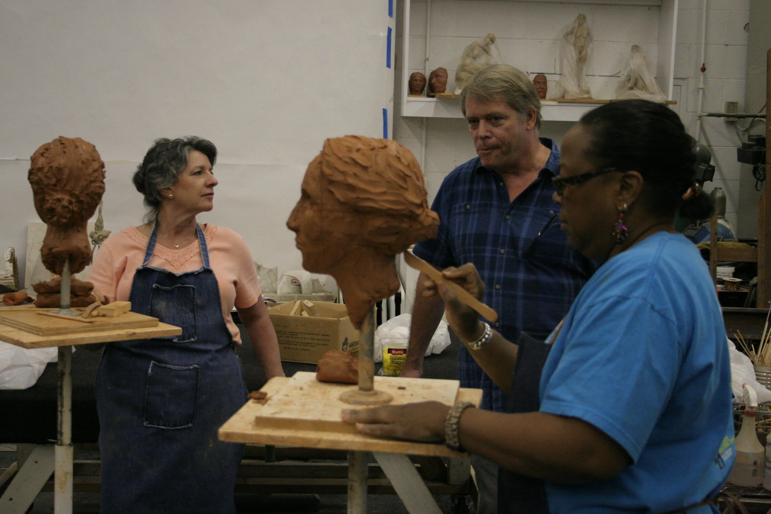 Three women and one man working in a ceramics studio with clay sculptures resembling large heads, with shelves of finished sculptures in the background.