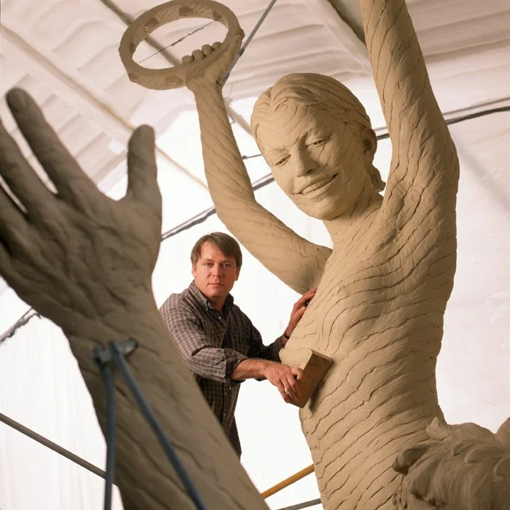 An artist working on a large clay sculpture of a woman with her arms raised, holding a hoop, while a man observes. The sculpture is in a studio.